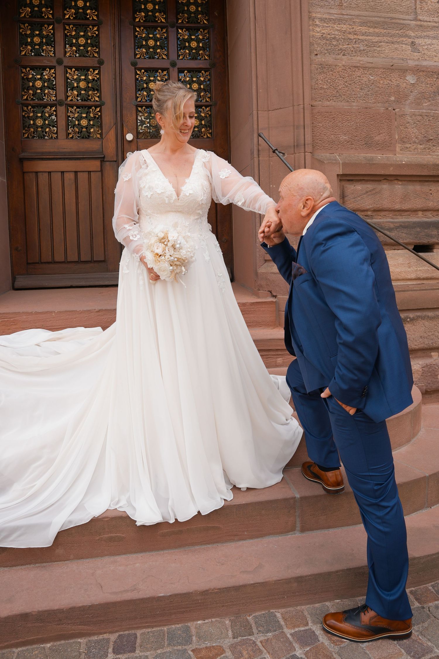 Groom in blue suit kissing bride's hand on church steps, bride wearing elegant white gown holding bouquet.