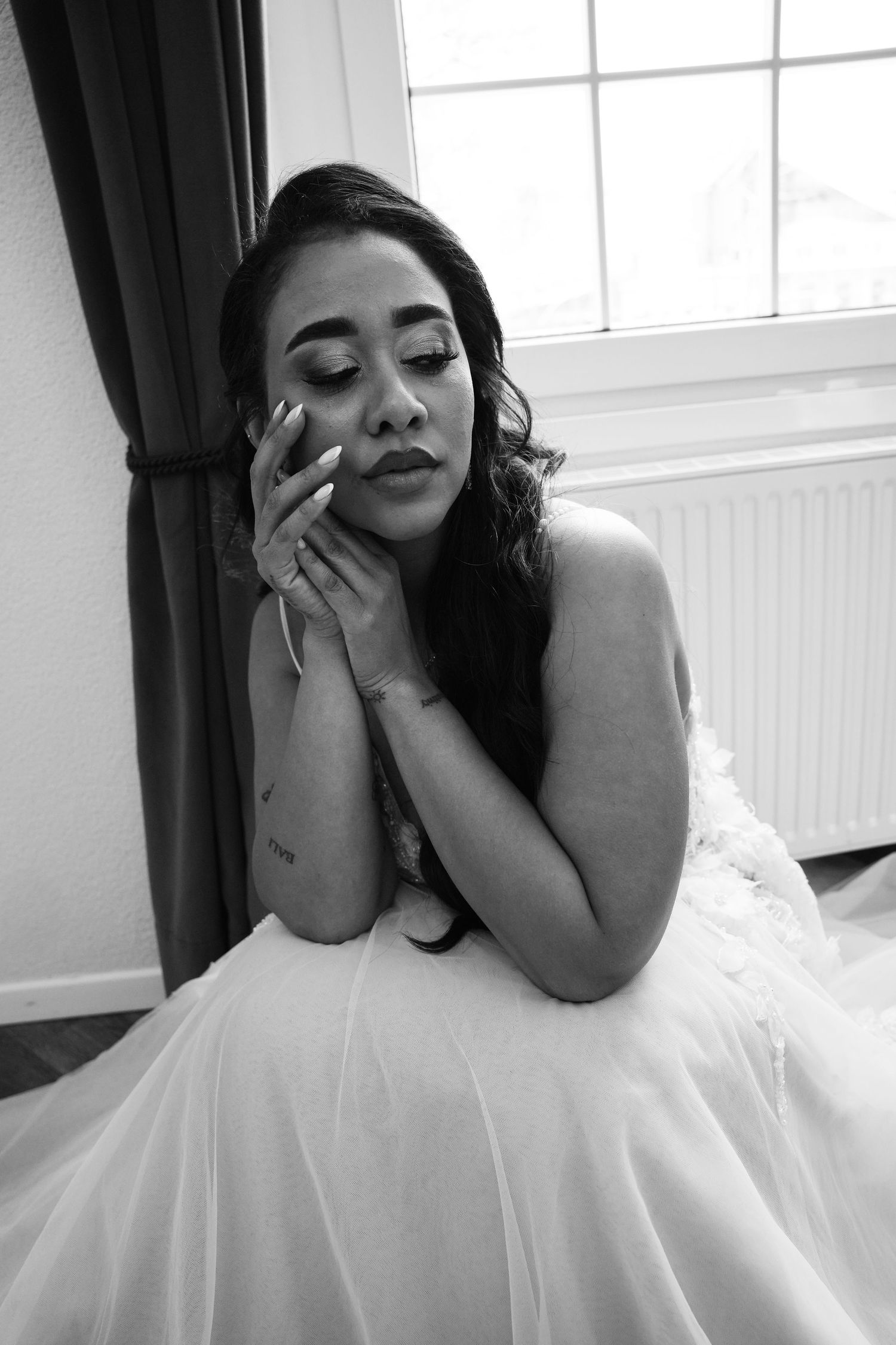Black and white portrait of a bride in a wedding gown, sitting by a window with eyes closed, hands gracefully touching her face.