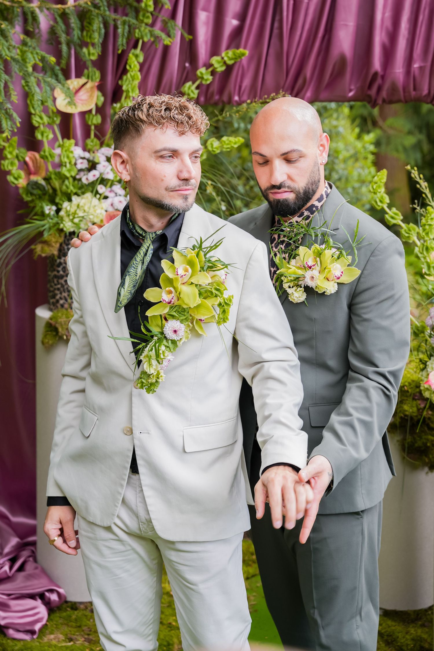Two grooms in suits with floral boutonnieres exchange rings at an outdoor wedding ceremony with purple draping.