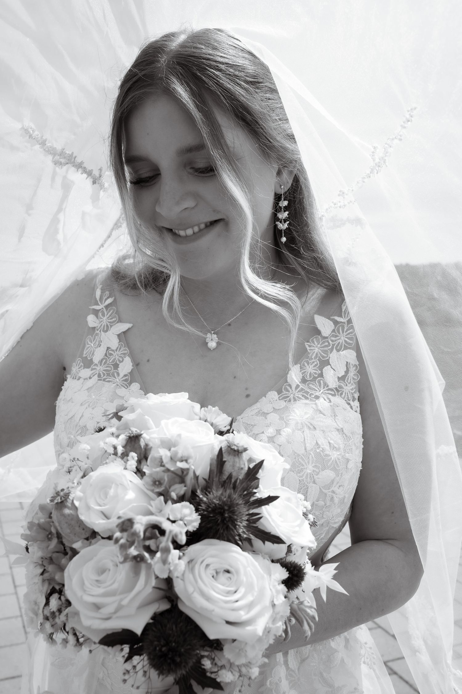 Black and white photo of a smiling bride in lace gown holding a bouquet of roses, wearing a veil and pendant necklace.