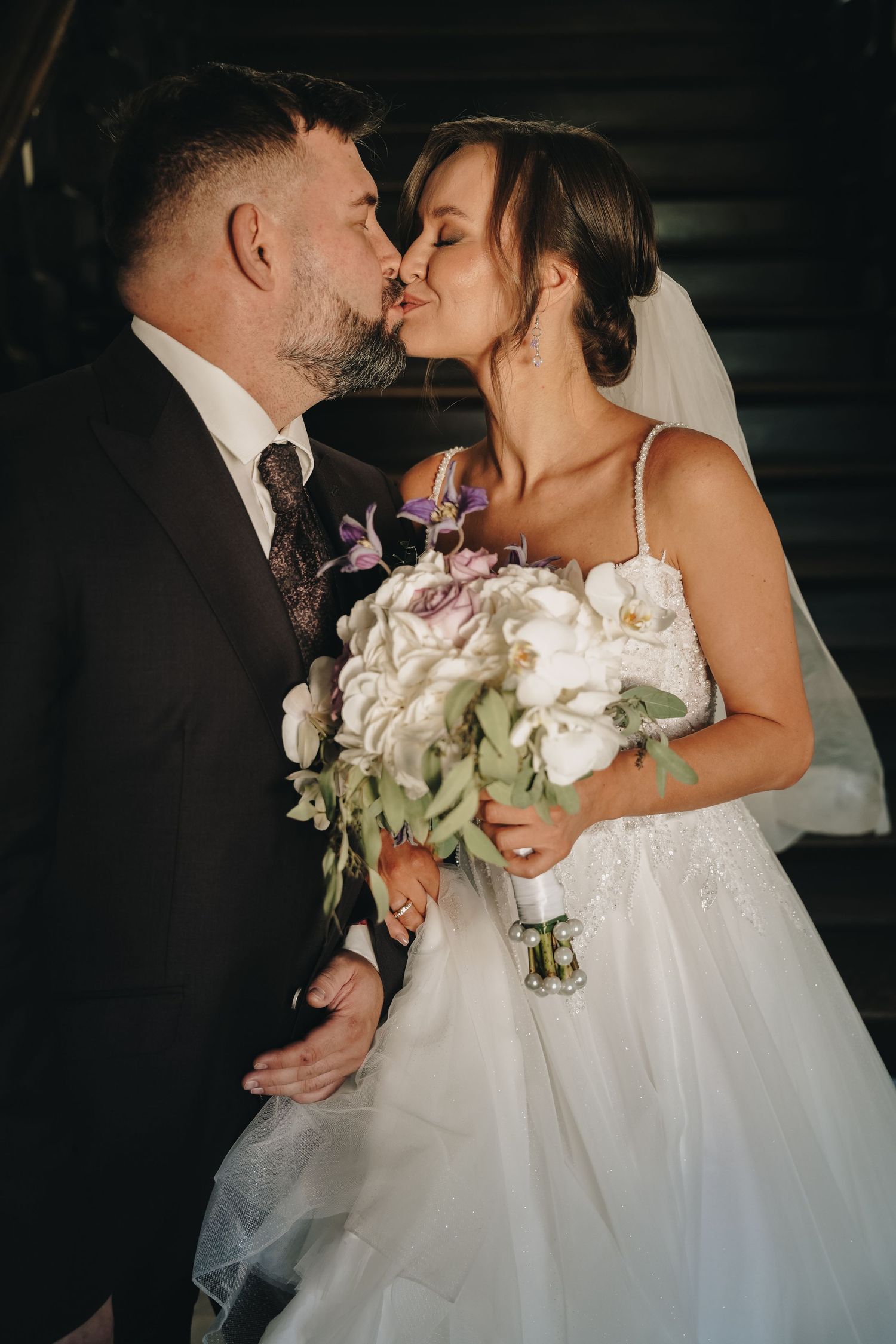 A bride and groom share a romantic kiss, she holds a white and purple floral bouquet, wearing a tulle gown with veil.