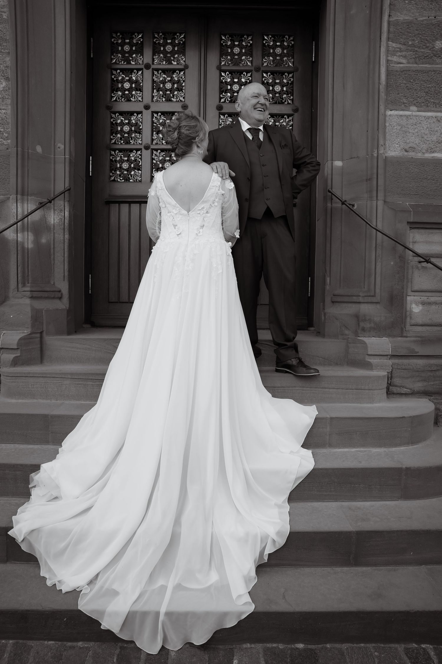 Black and white photo of a bride in a flowing white gown with train, standing with a groom on church steps.