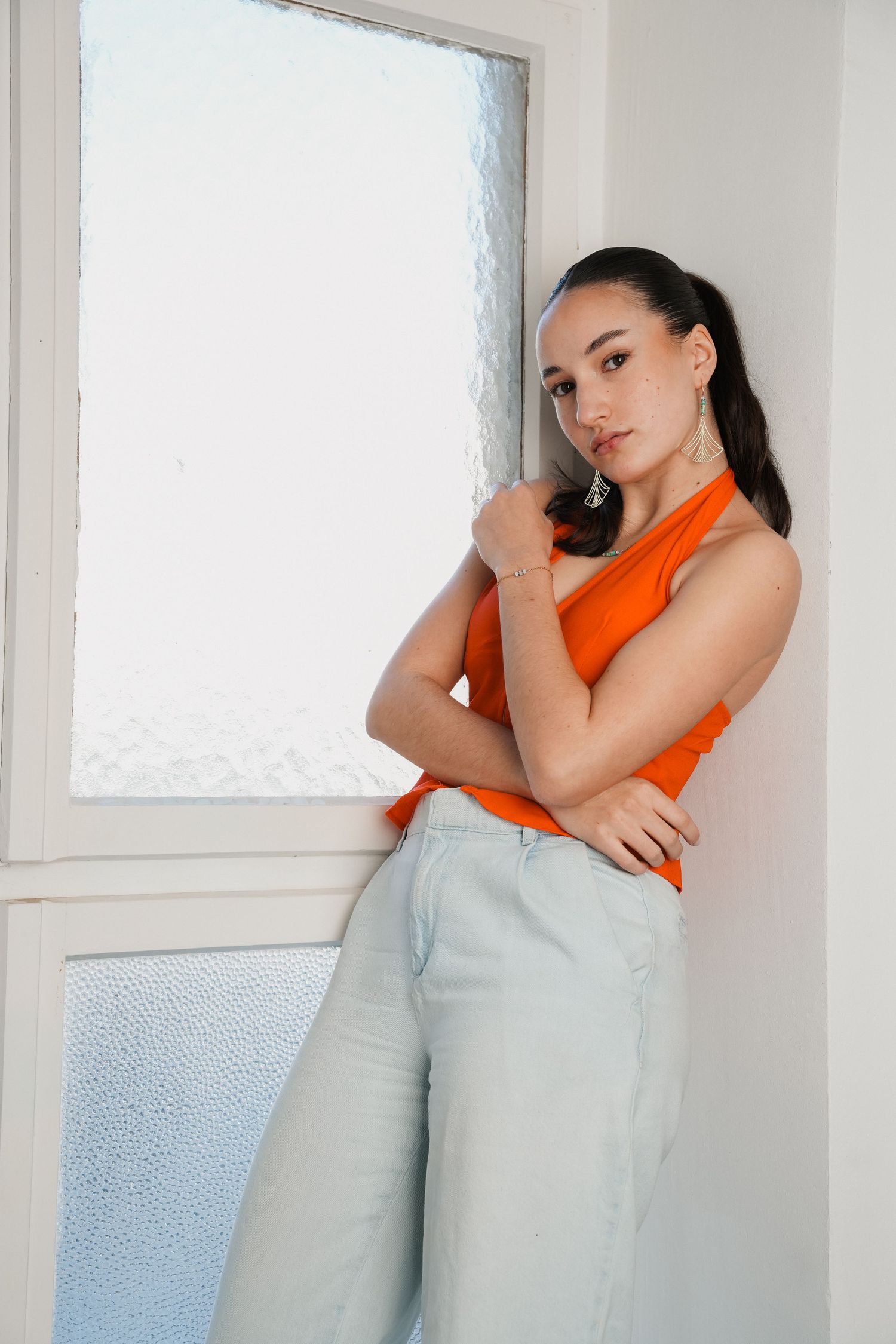 Young woman in orange top and light jeans posing by a frosted glass window in a bright white room.