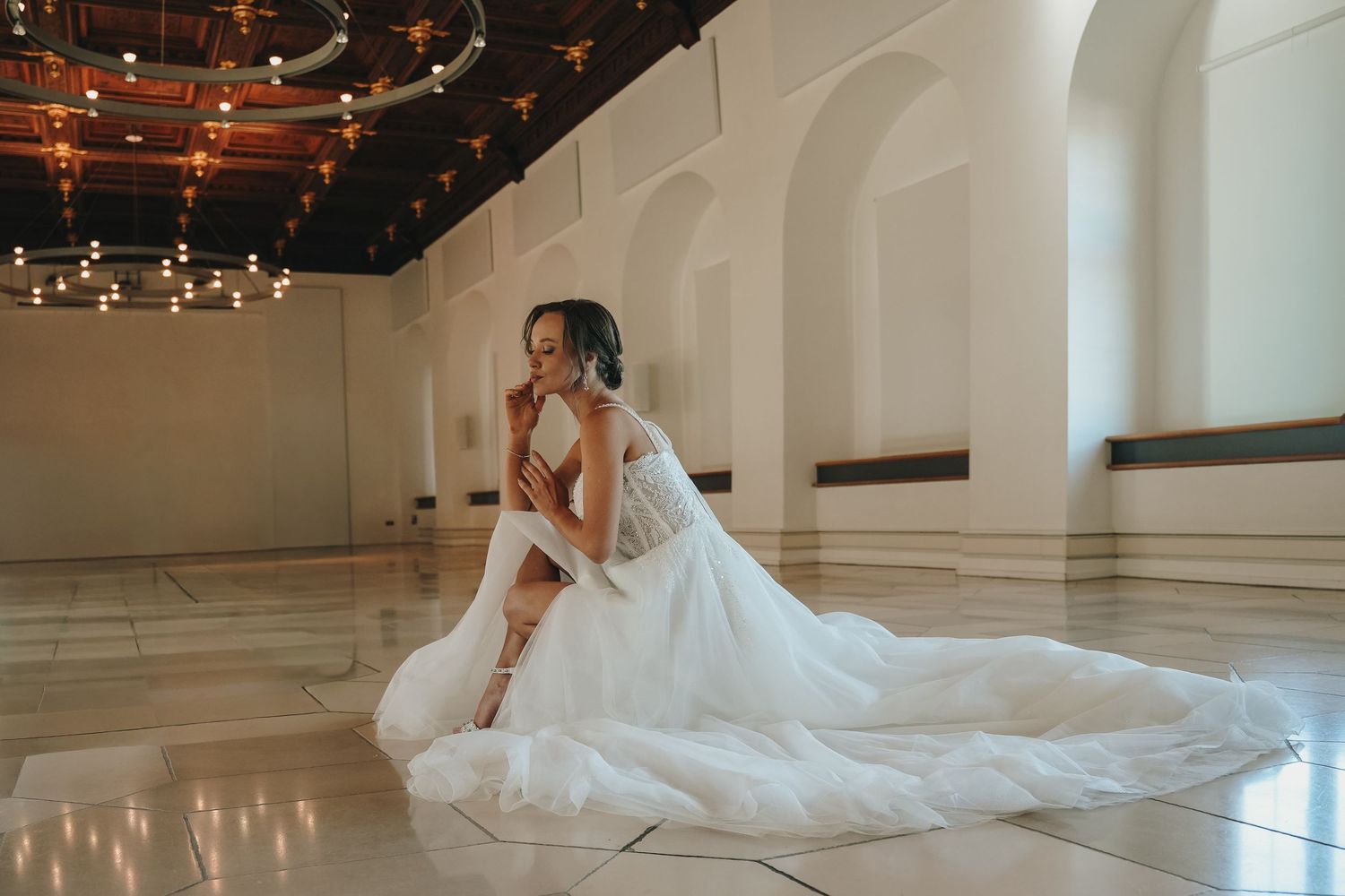 Bride in a flowing white wedding gown sits gracefully on marble floor in an elegant ballroom with chandeliers.
