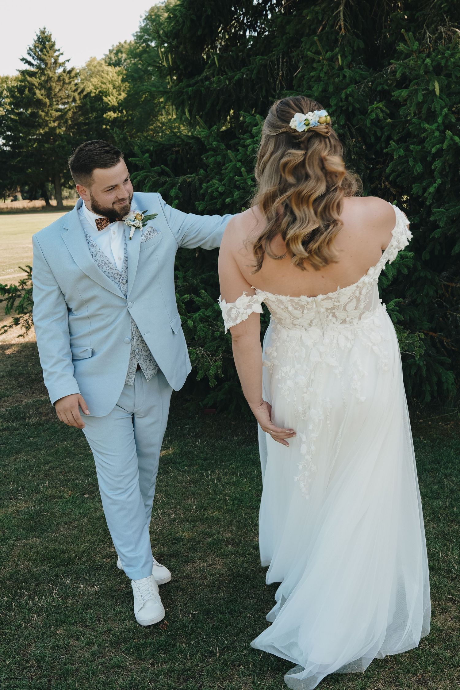 Groom in light blue suit with bow tie smiles at bride in off-shoulder lace wedding dress outdoors.