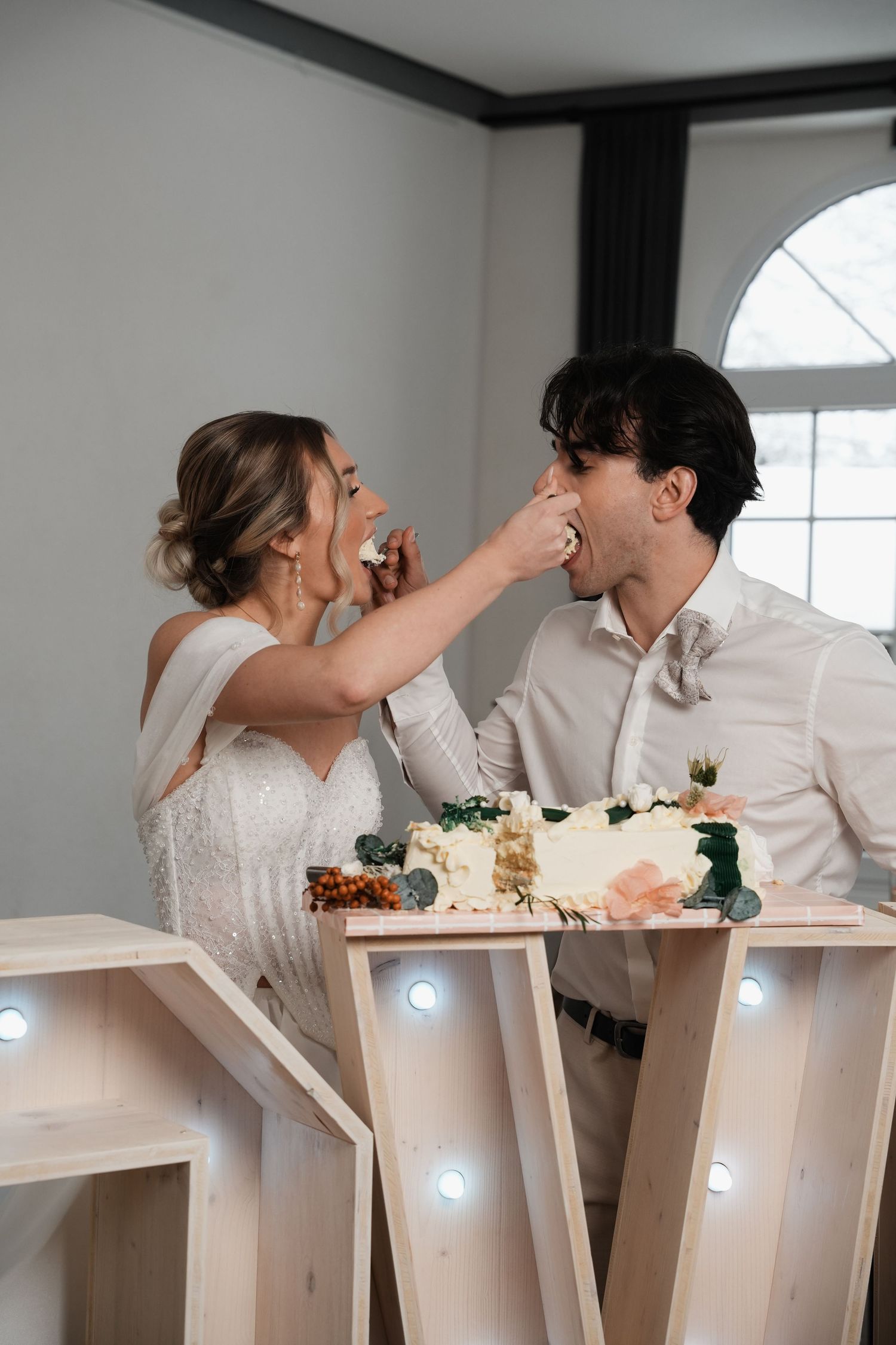 Bride and groom feeding each other wedding cake beside illuminated LOVE letters display.