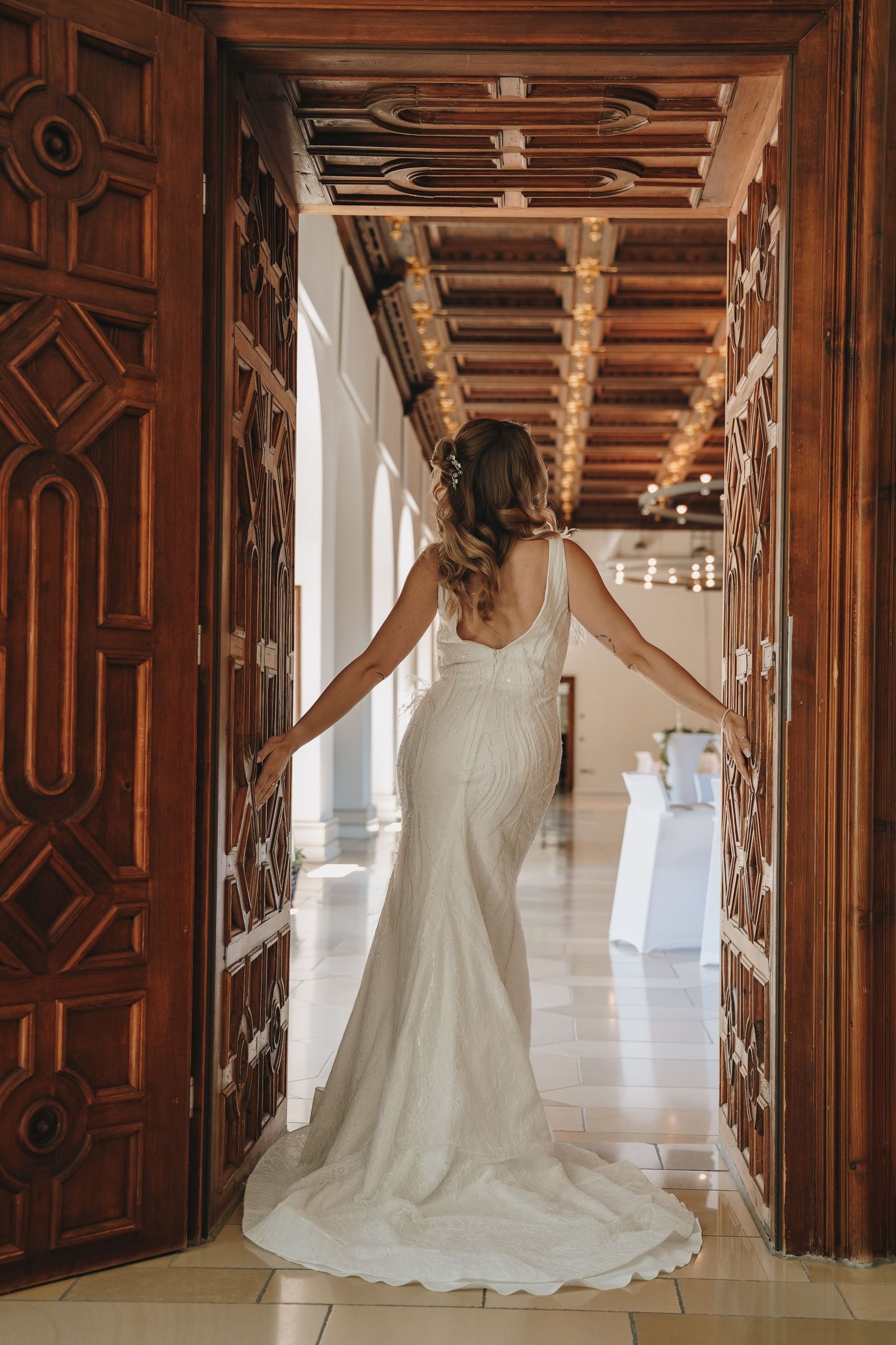 Bride in elegant white mermaid gown stands between ornate wooden doors in a luxurious venue hallway.