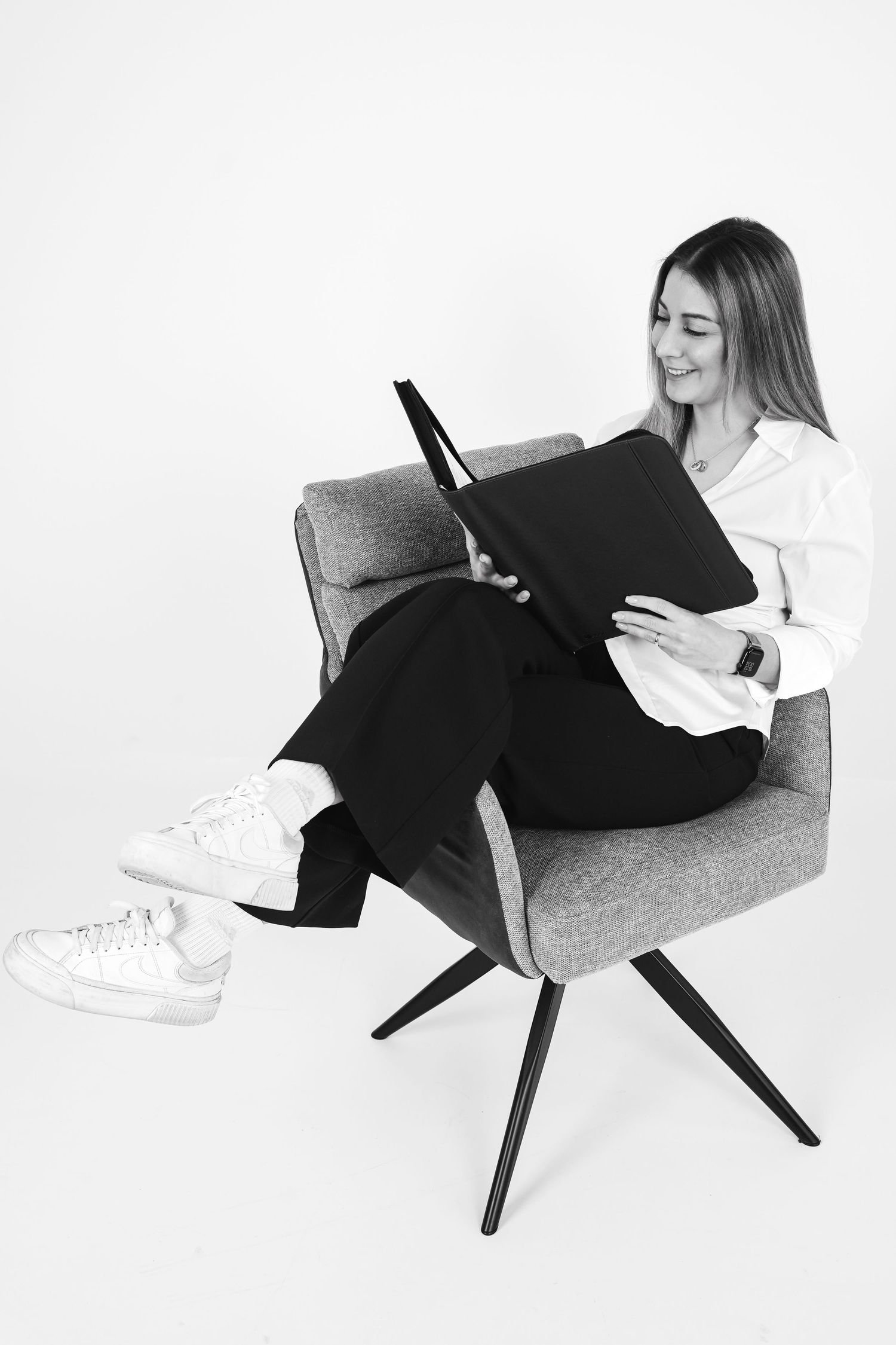 Young woman sitting in gray chair smiling at tablet, wearing white blouse and black pants with white sneakers.