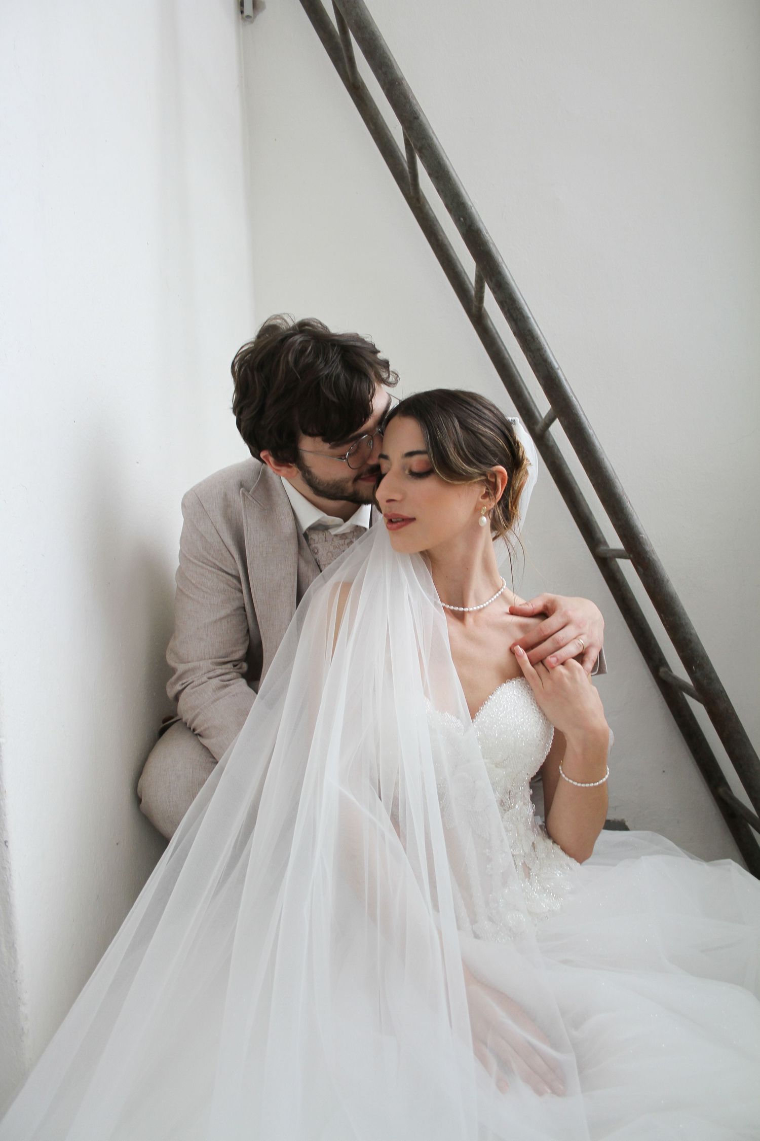 A groom in a gray suit tenderly kisses his bride wearing a white lace gown and veil on a staircase.