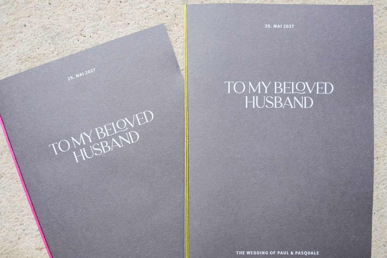 Two gray wedding vow booklets titled To My Beloved Husband with pink and yellow spine accents on a marble surface.