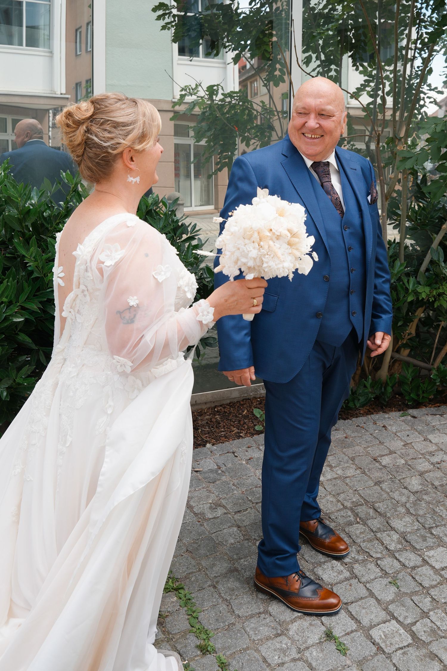 Bride in white gown holding bouquet with smiling groom in blue suit on cobblestone path outdoors.