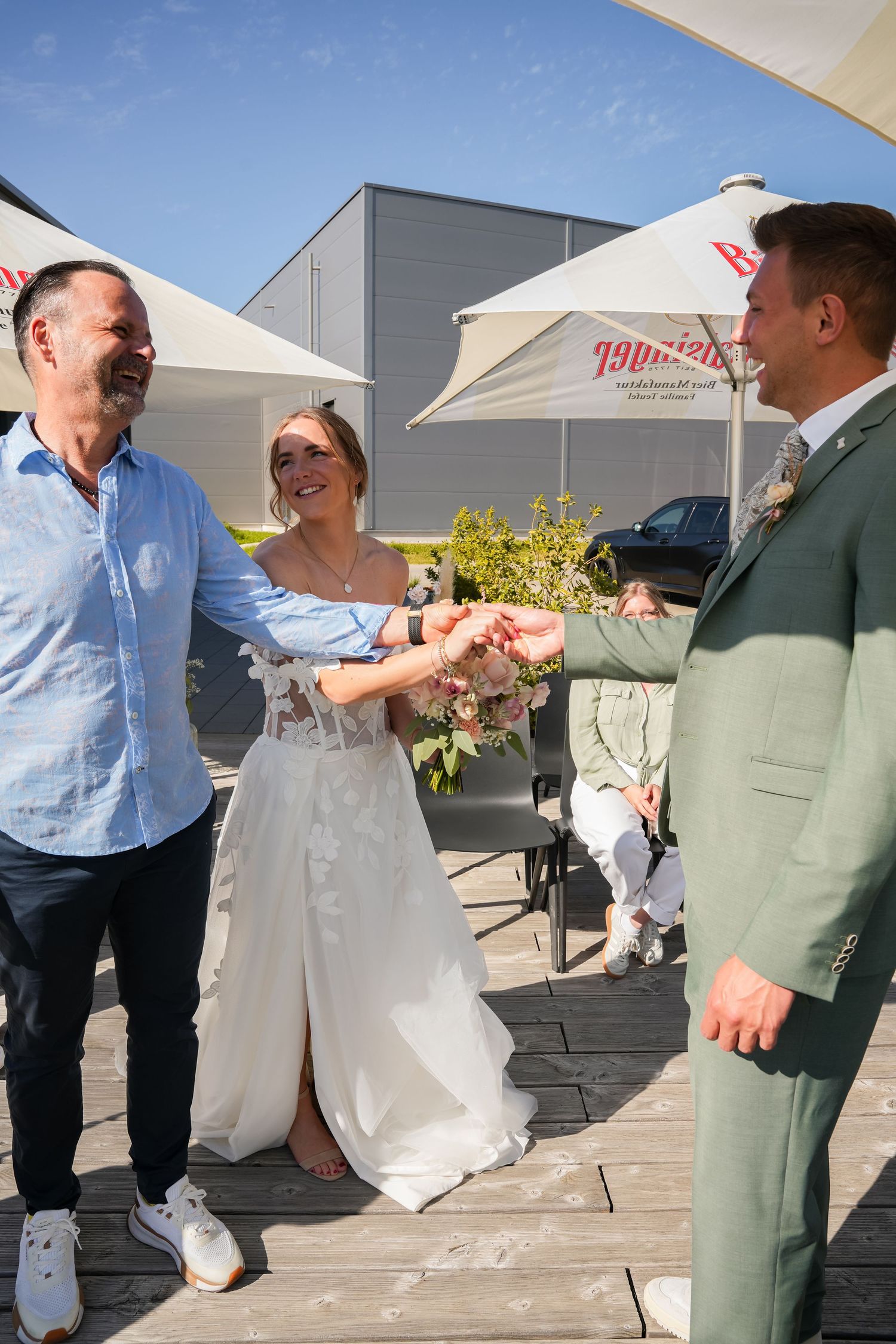 Bride in white dress stands between groom in green suit and guest shaking hands outdoors at sunny wedding reception.