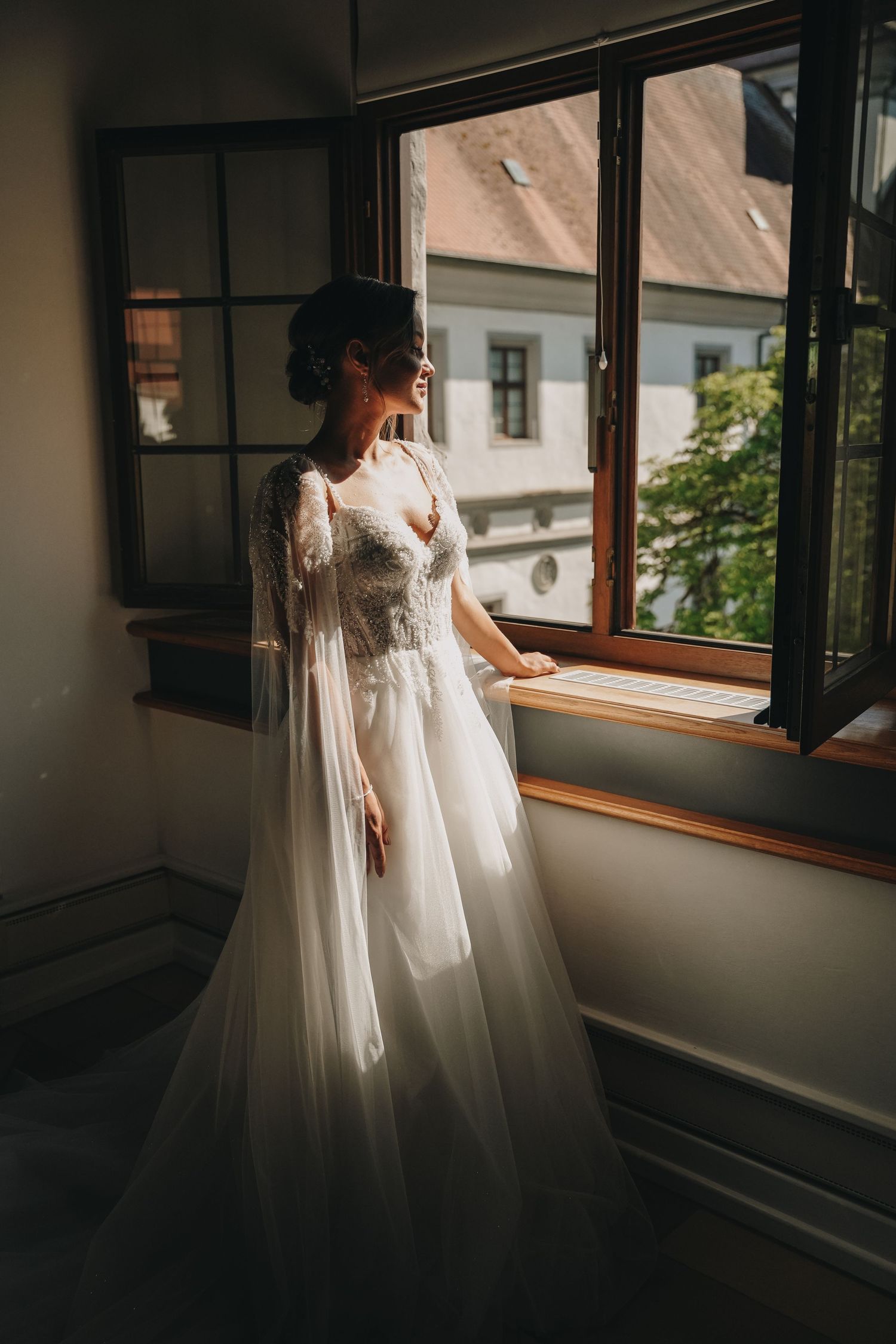 Bride in elegant white tulle wedding gown with lace bodice standing by open window in soft natural light.