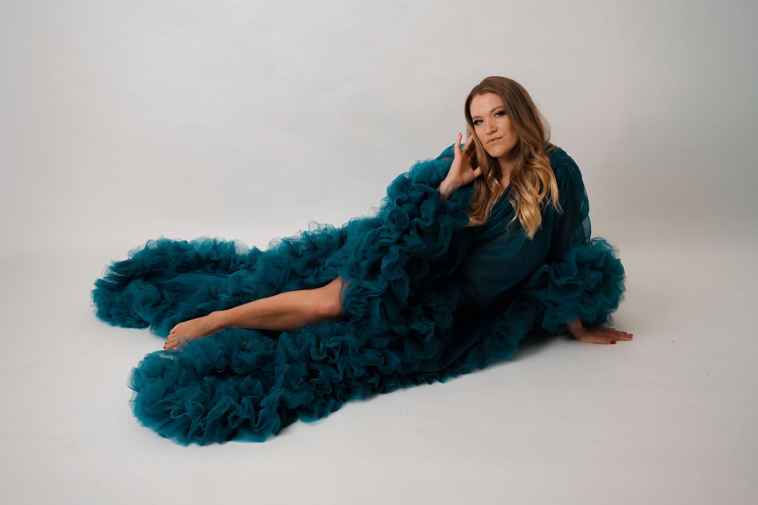 Woman in teal ruffled tulle gown posing elegantly on white studio floor.