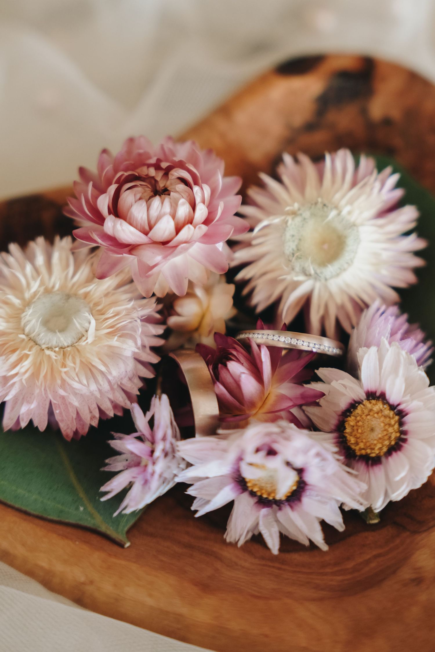 Pink and white dried flowers with wedding rings in a rustic wooden bowl.
