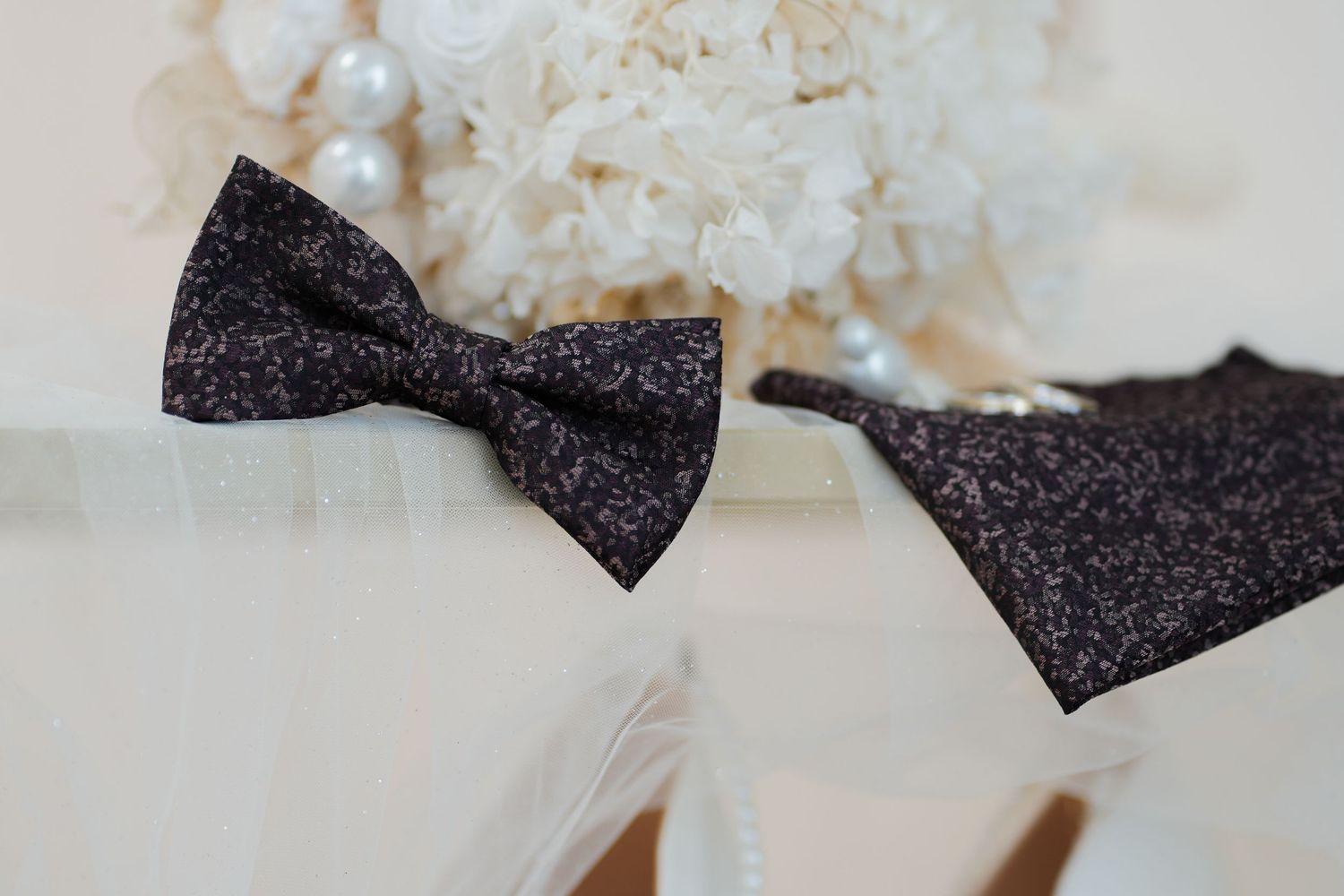 Black glitter bow tie and pocket square displayed beside a white floral bouquet with pearl accents.
