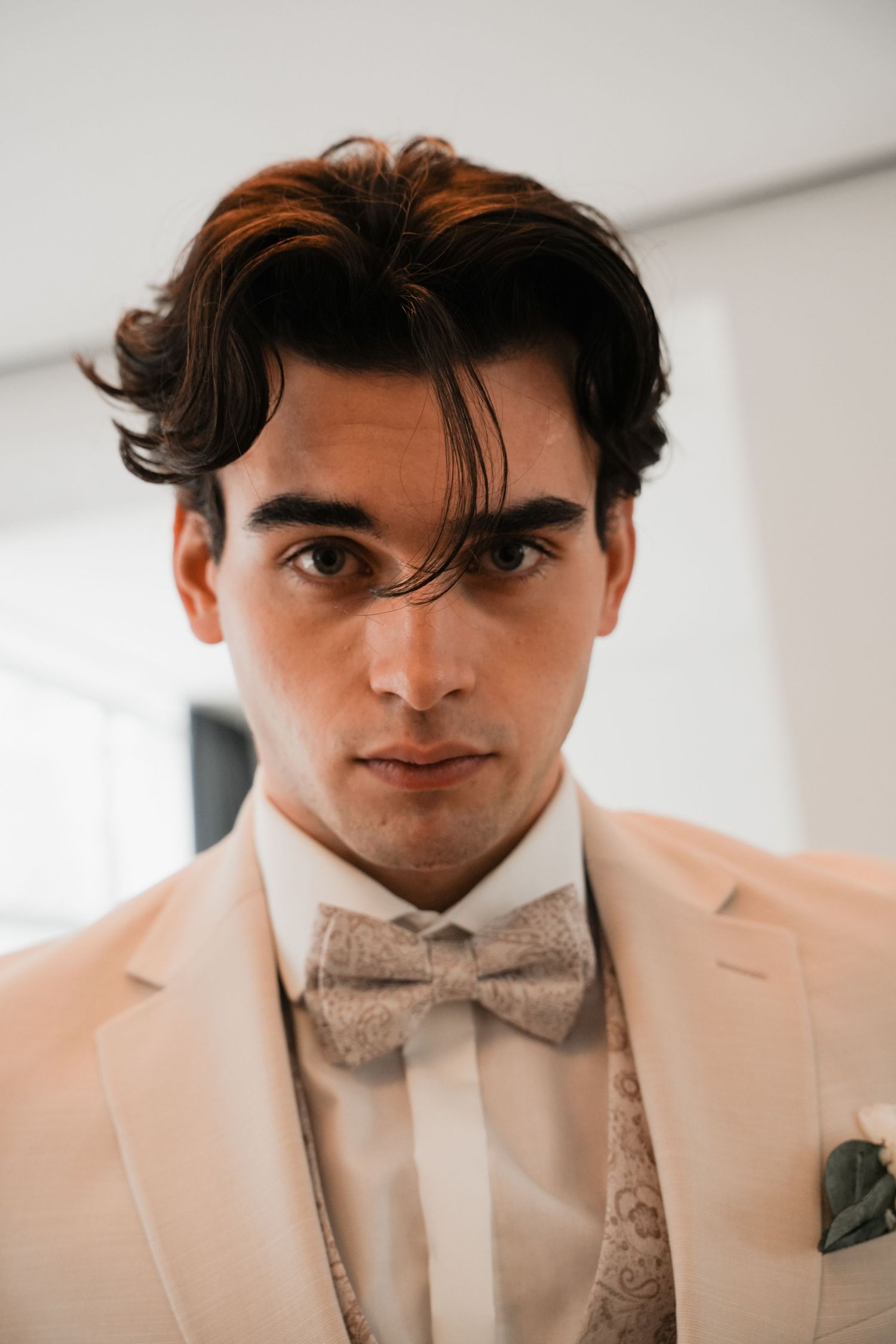 Young man in cream suit with paisley bow tie and pocket square, styled dark wavy hair, serious expression.