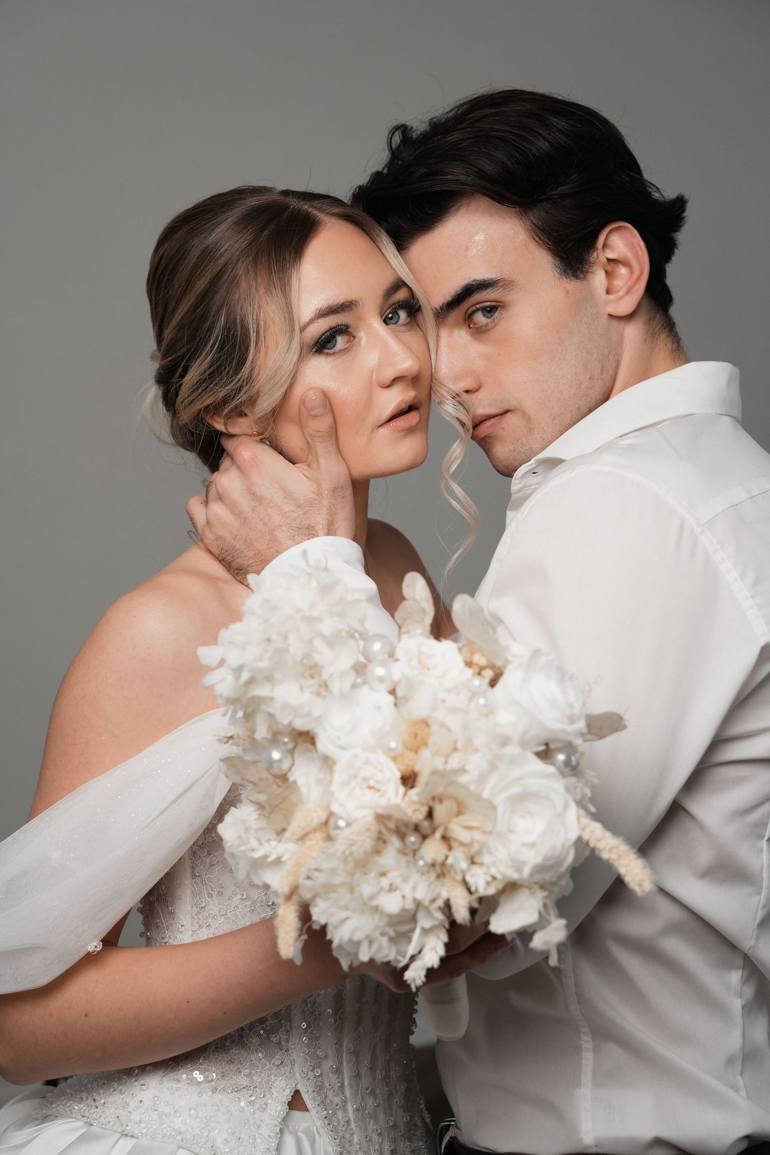 Bride and groom posing closely together, holding a white floral bouquet, in elegant wedding attire against a gray background.