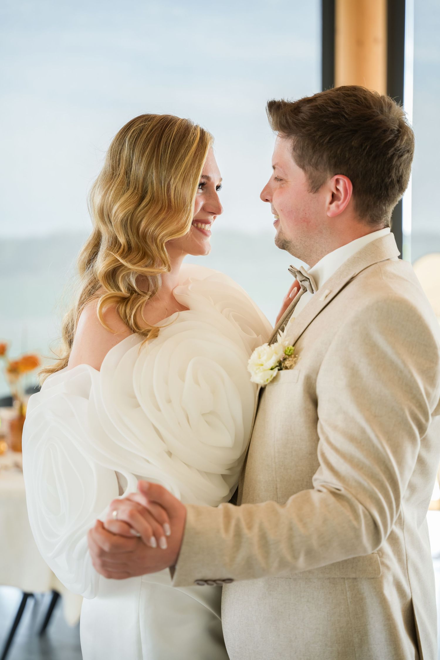 Bride and groom share first dance, she in white gown, he in beige suit with boutonniere, ocean view backdrop.
