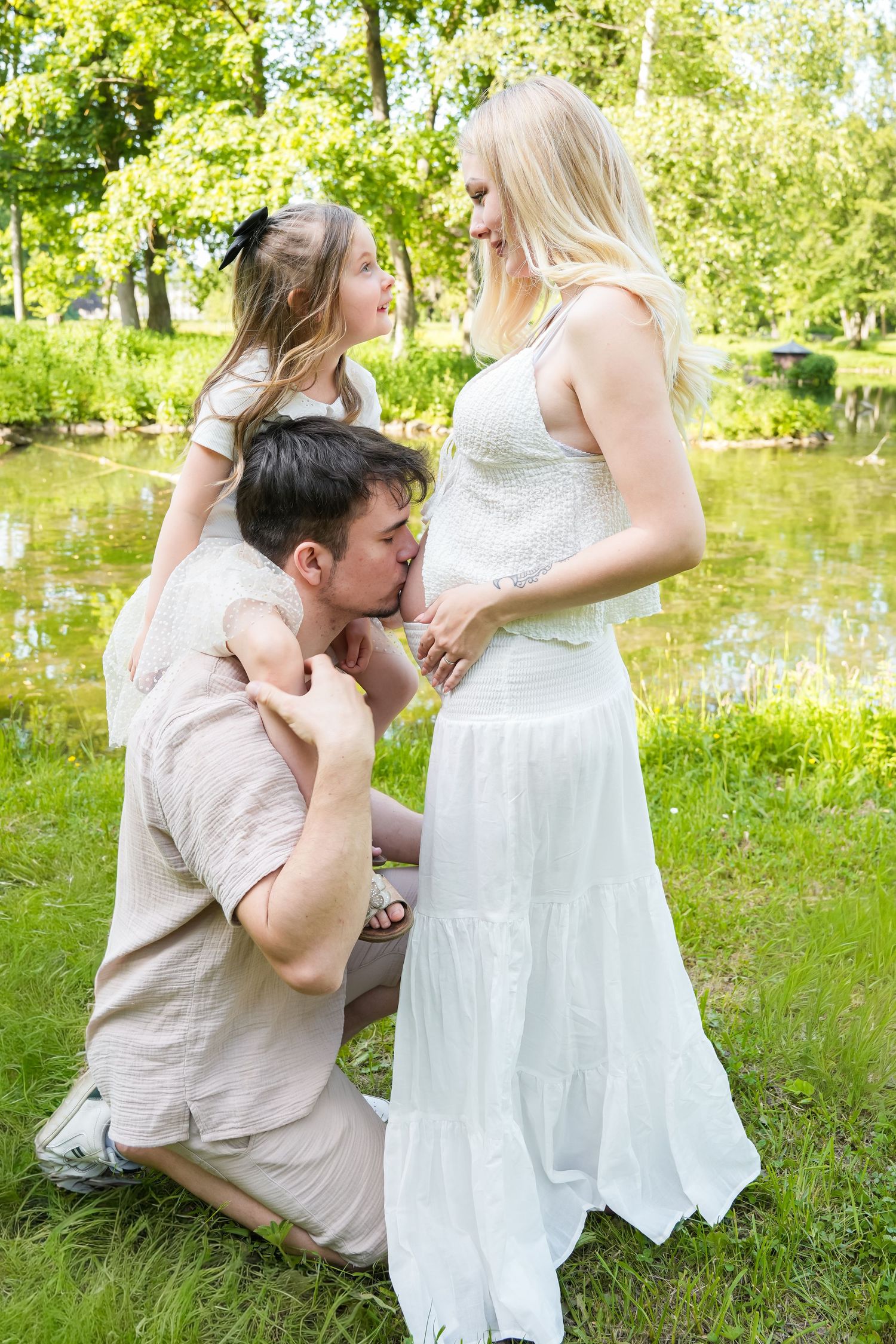 A man kneels kissing a pregnant woman's belly while a young girl sits on his shoulders in a sunny park.