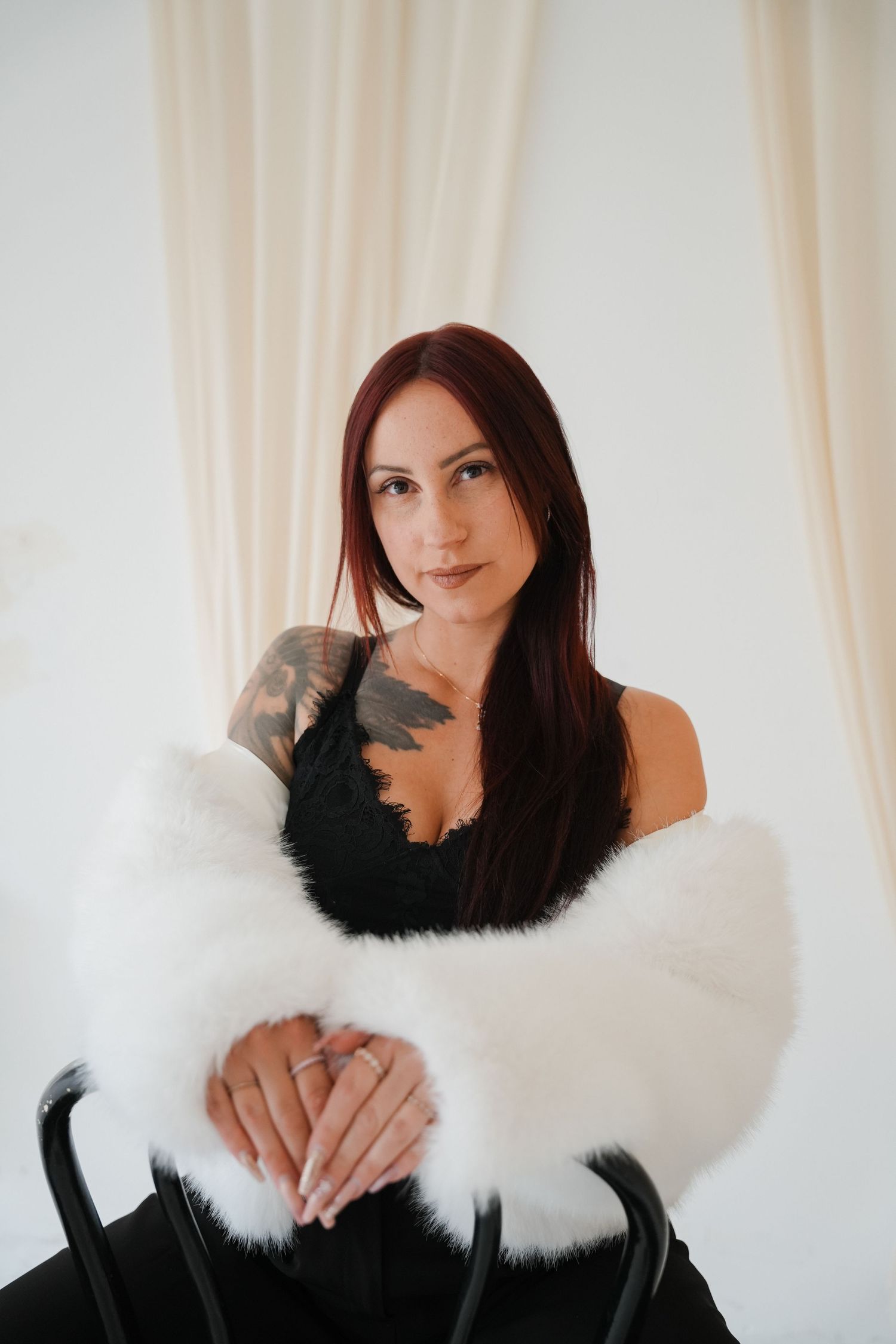 Woman with red hair and tattoos wearing black lace top and white fur wrap, seated elegantly in a bright studio.