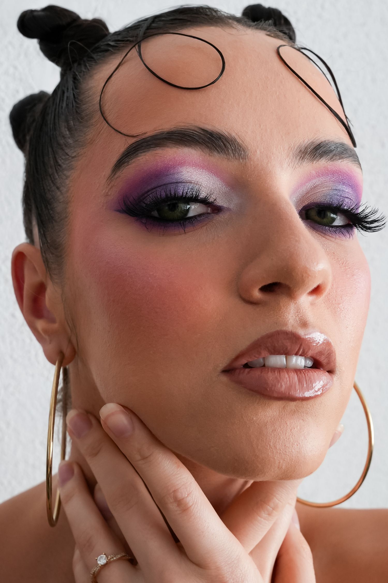 Close-up of a woman with bold purple eyeshadow, dramatic lashes, rosy blush, glossy lips, and gold hoop earrings.