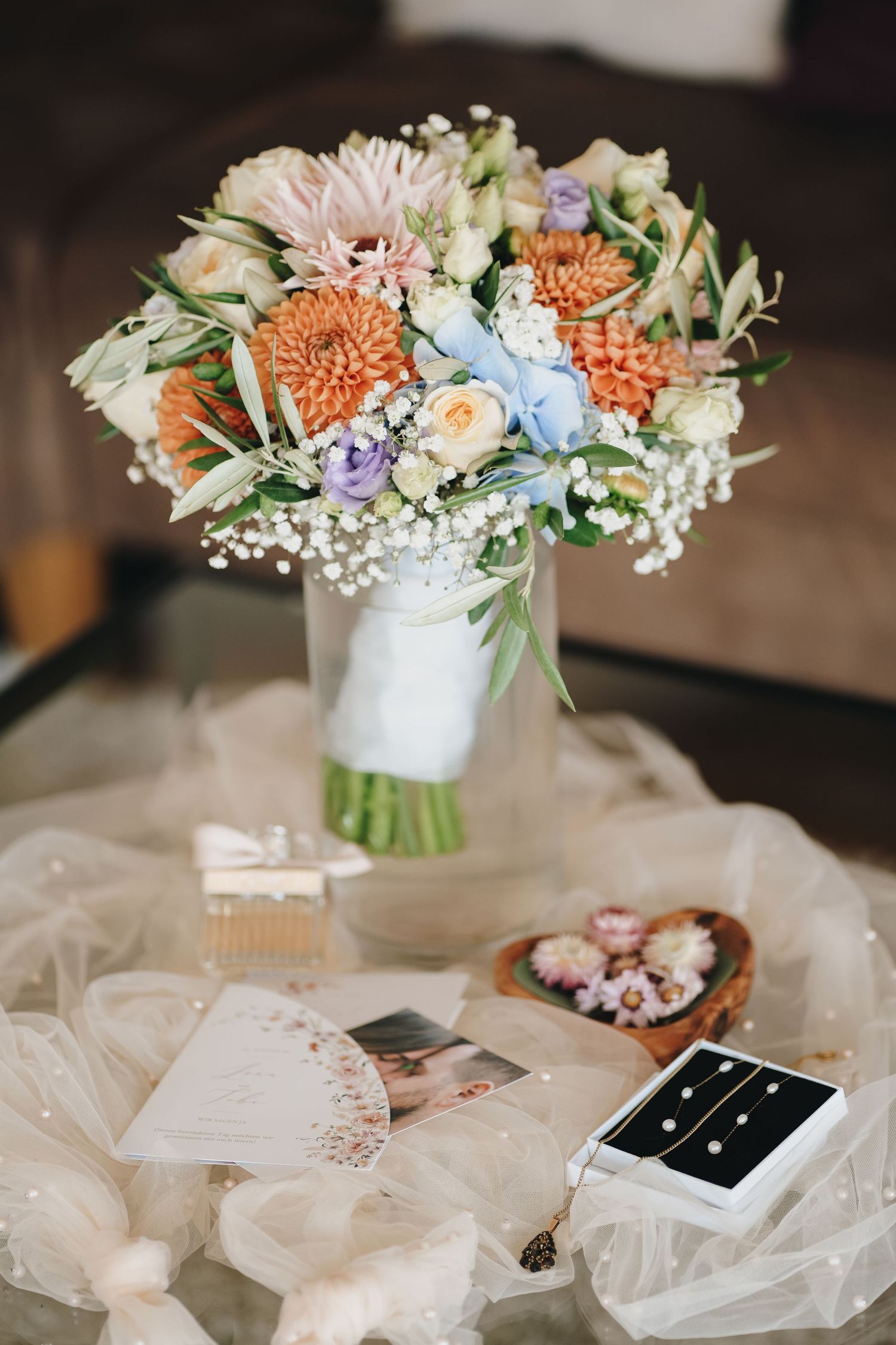Pastel wedding bouquet of orange, peach and lavender flowers with baby's breath in a glass vase beside a paper fan and jewelry box.
