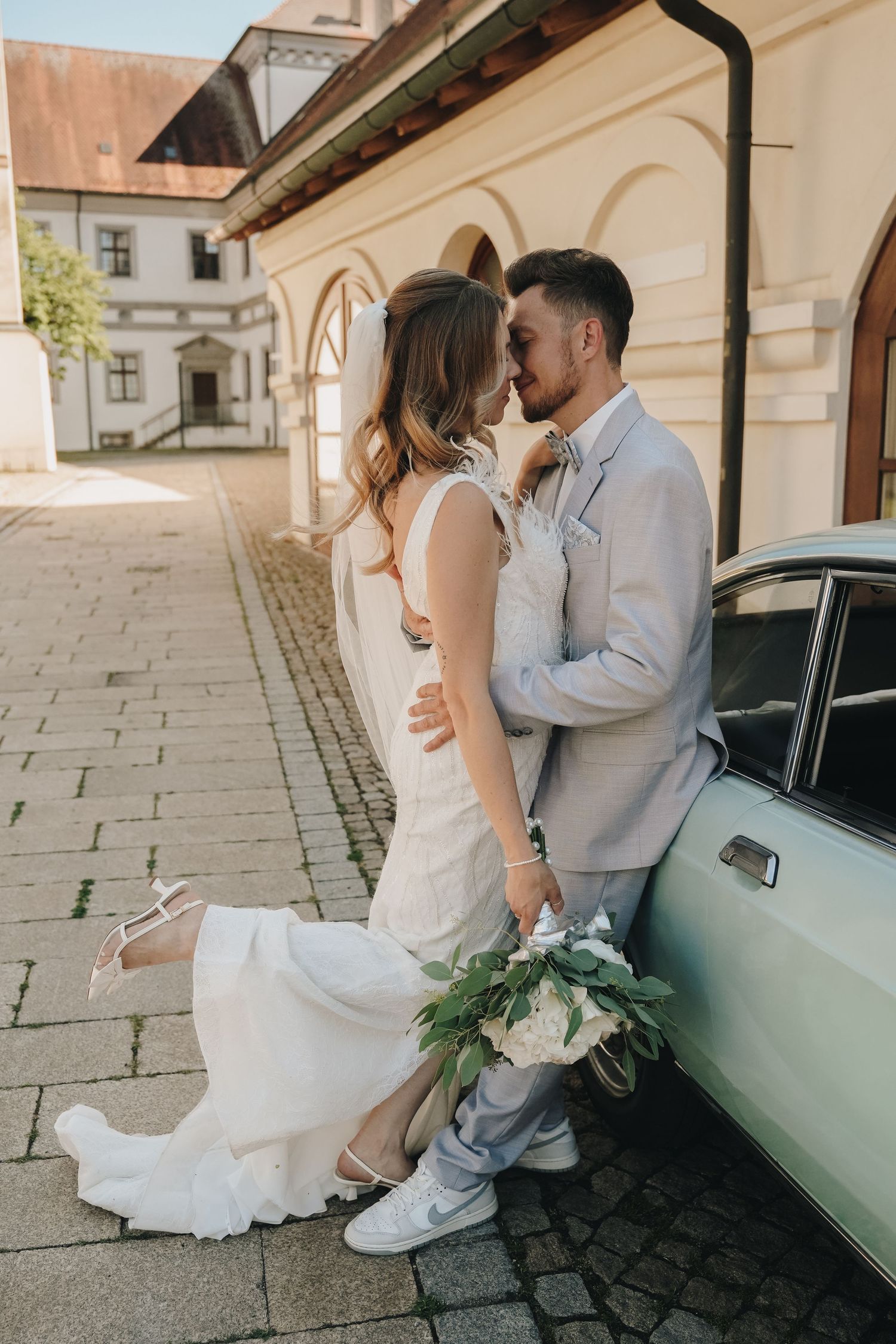 Bride and groom sharing a romantic moment by a vintage car, bride in white dress holding bouquet, groom in light gray suit.