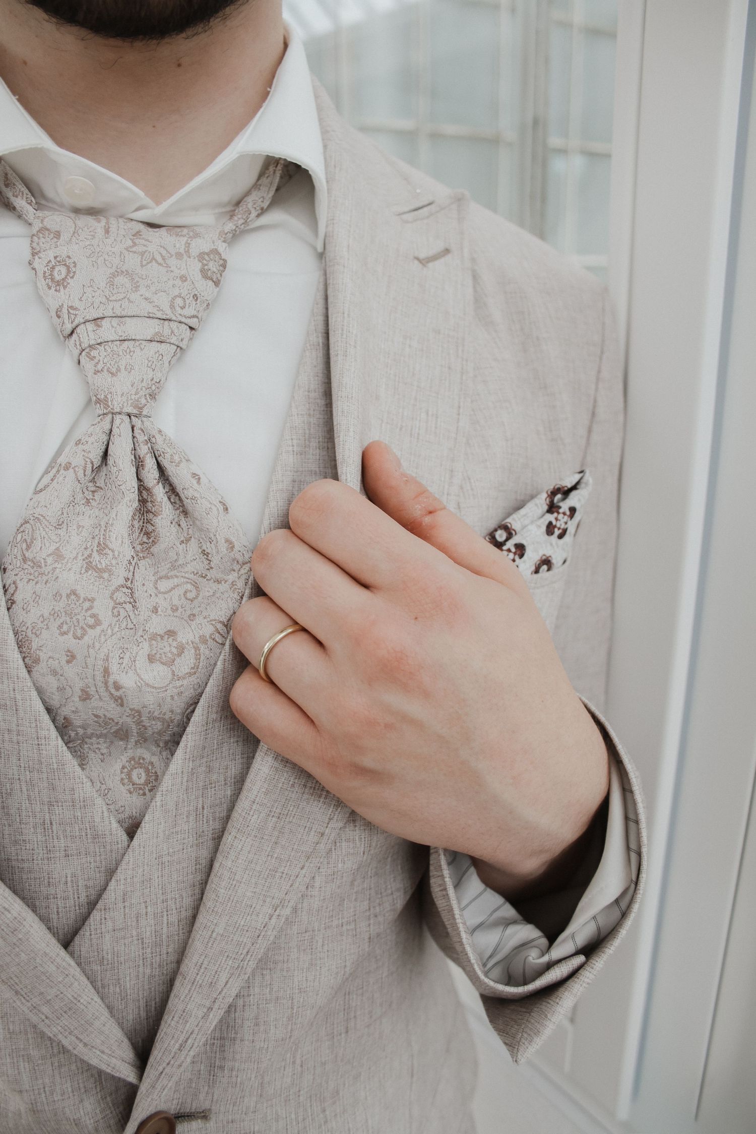 Groom in a beige three-piece suit with paisley tie and pocket square, wearing a gold wedding band.