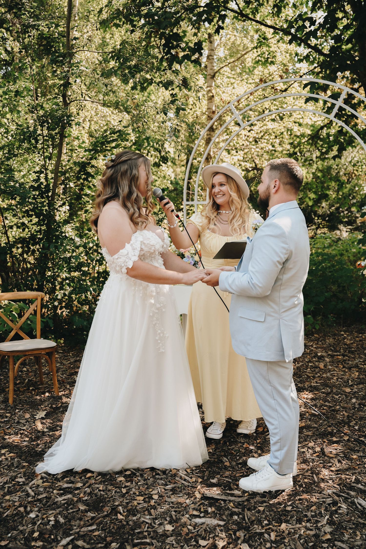 Bride and groom exchange vows outdoors in a lush garden ceremony, officiated by a woman in a yellow dress.