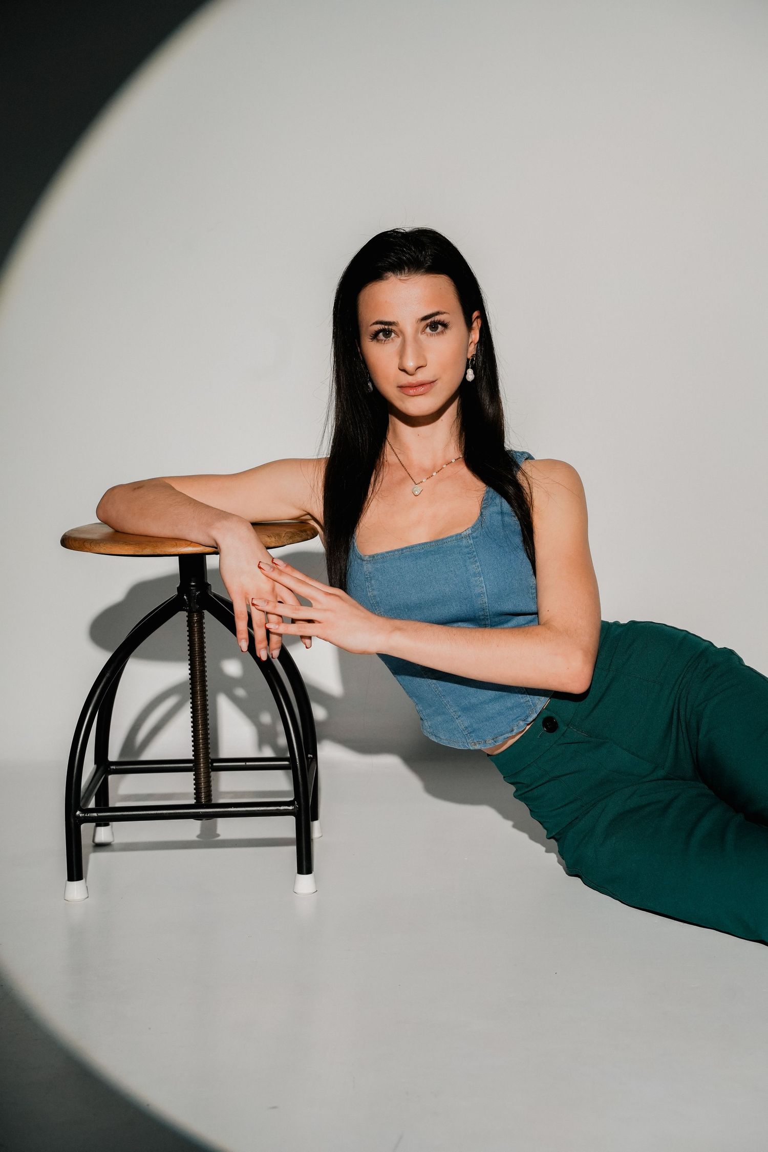 Woman in denim top and green pants posing beside a wooden stool against a white studio background.