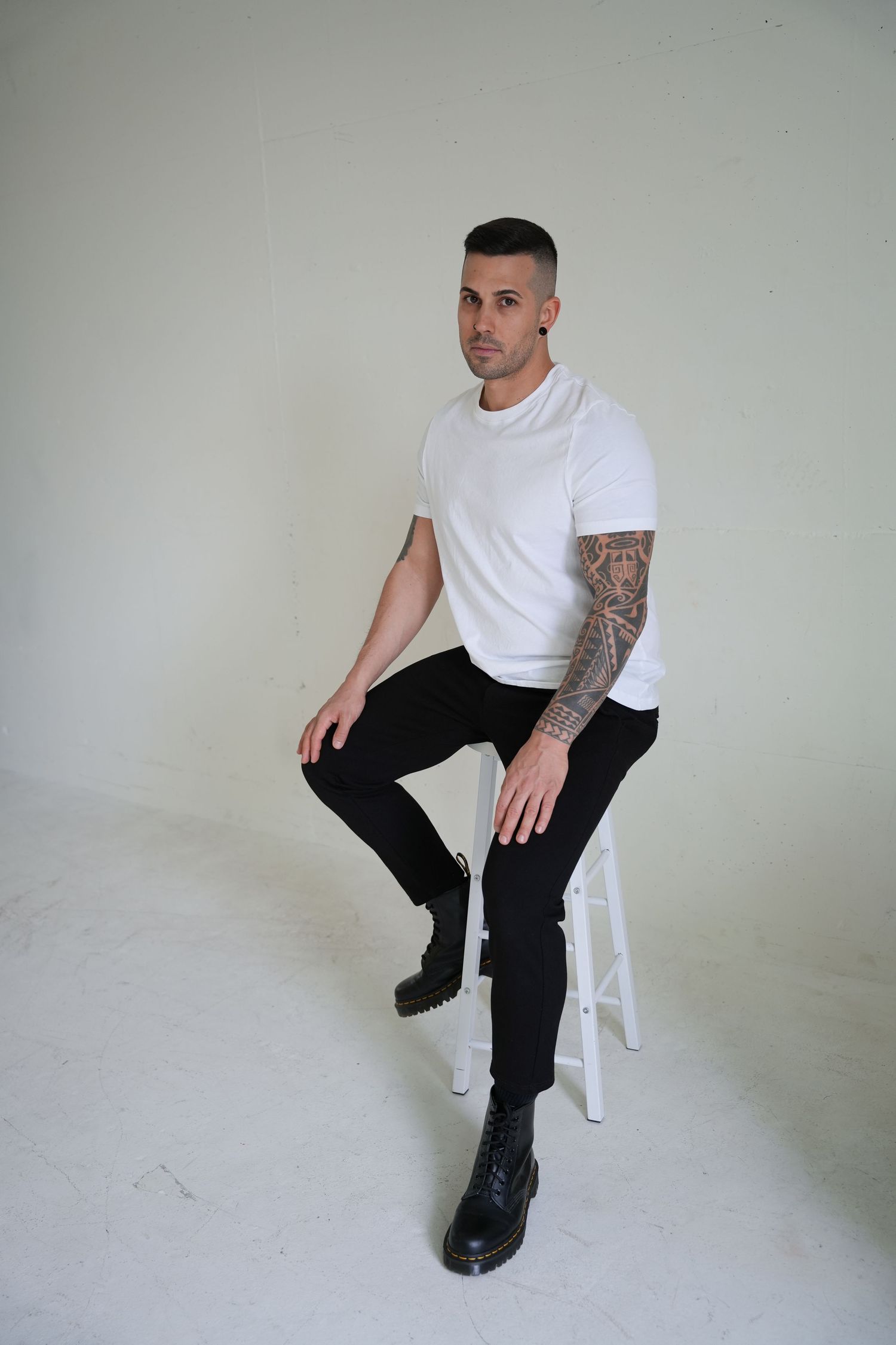 Tattooed man in white t-shirt and black jeans sitting on a stool wearing black boots against white background.