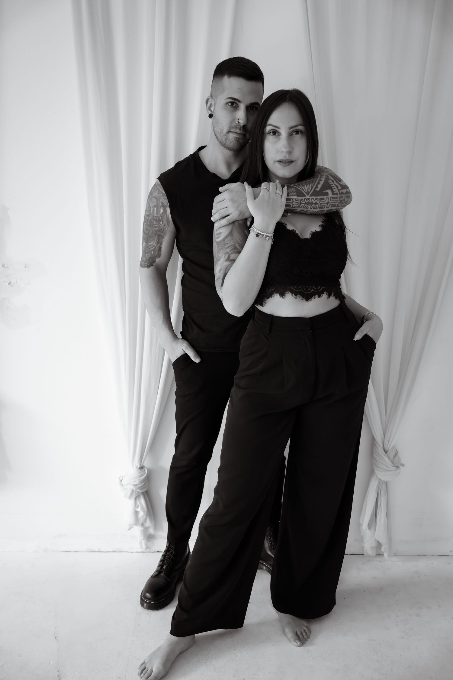 Black and white photo of a couple in matching black outfits, man with tattoos embracing woman from behind.