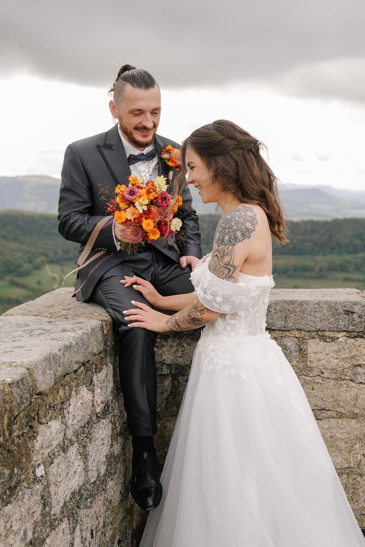 Smiling bride in a tattooed off-shoulder lace gown sits beside a groom holding an orange bouquet on a stone wall above green hills.