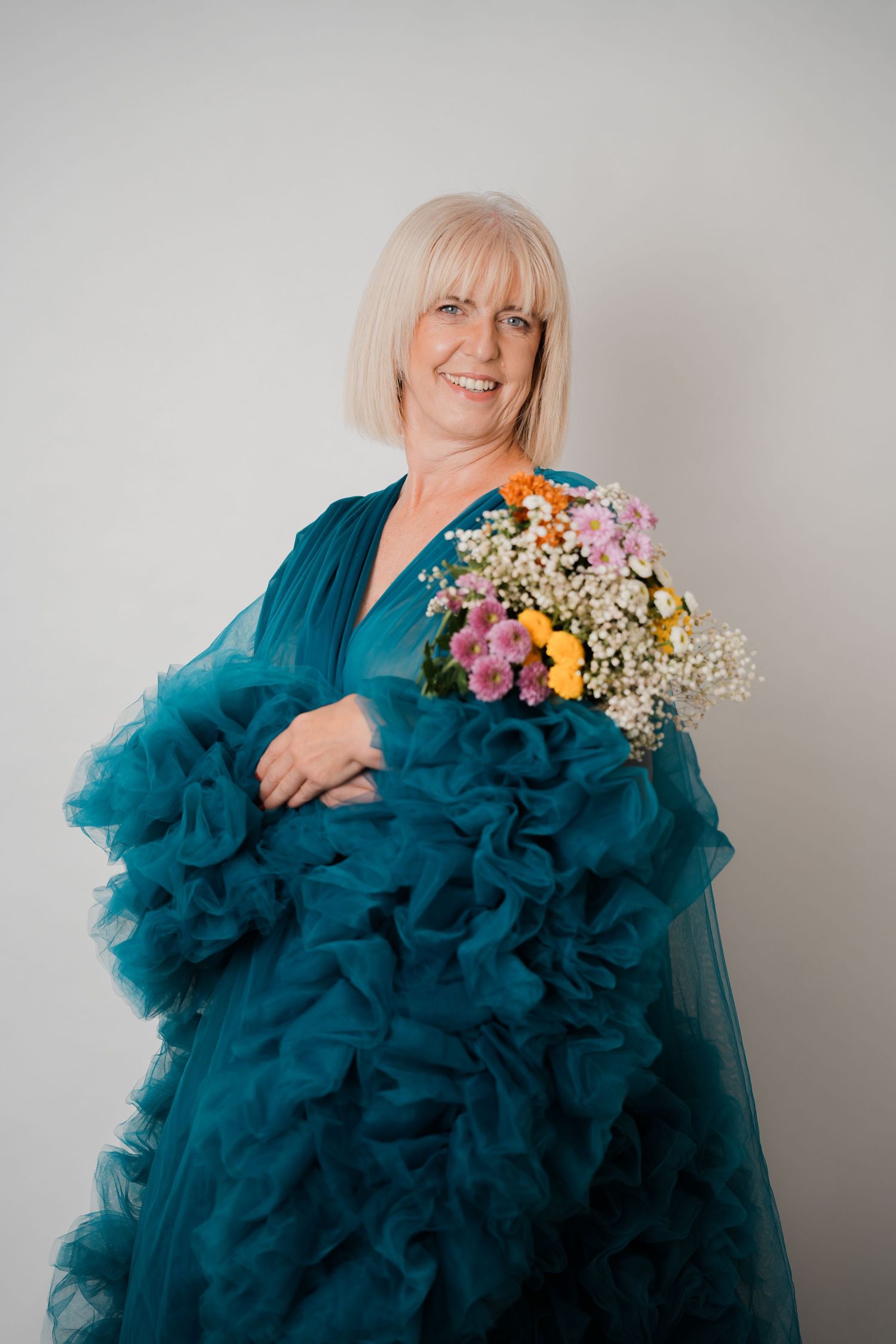 Smiling older woman with blonde bob wearing a teal ruffled gown, holding a colorful bouquet of wildflowers.