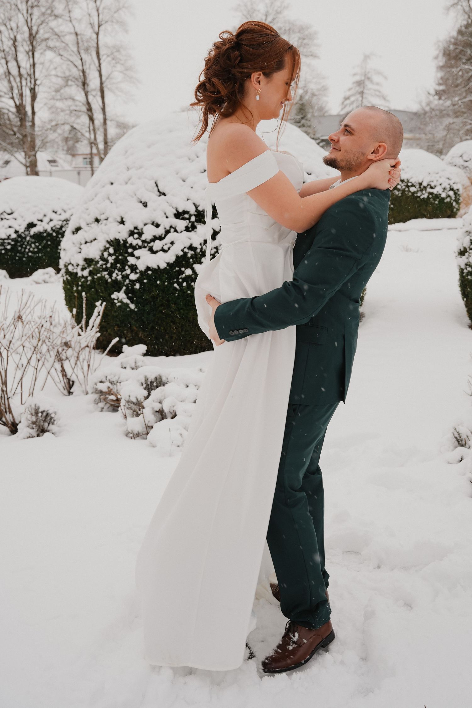 Bride and groom embracing in snowy winter garden, she in white gown, he in green suit.