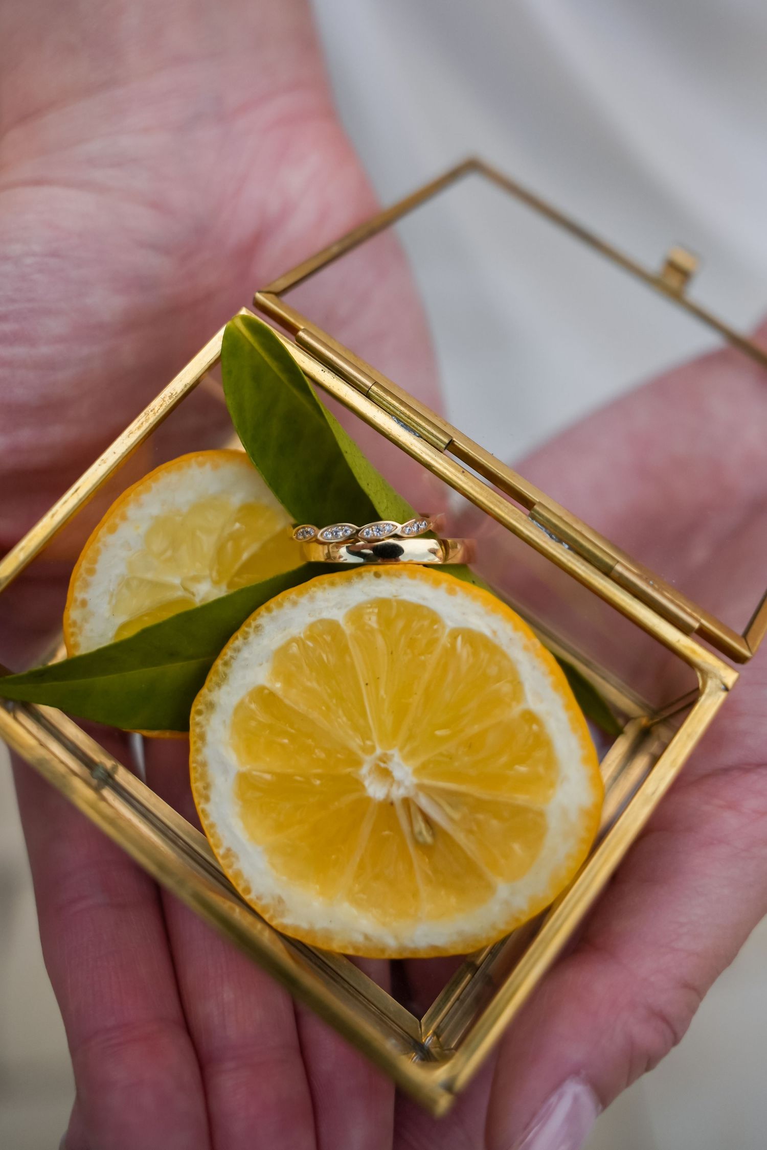 Hands holding a gold glass box with lemon slices, green leaves, and a diamond wedding ring inside.