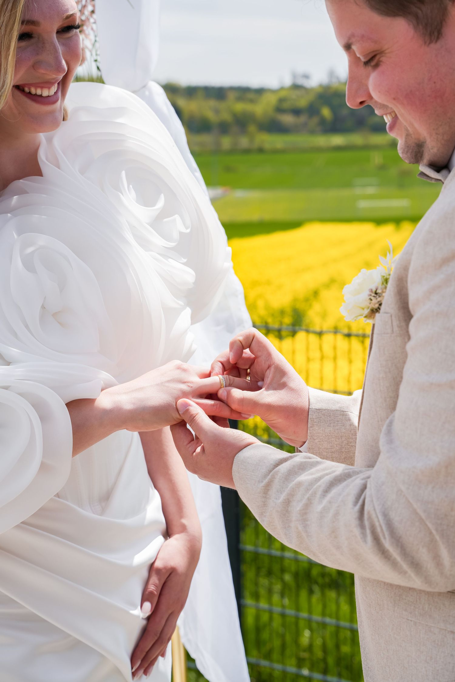 Groom placing wedding ring on bride's finger outdoors, with yellow flower fields in the background.