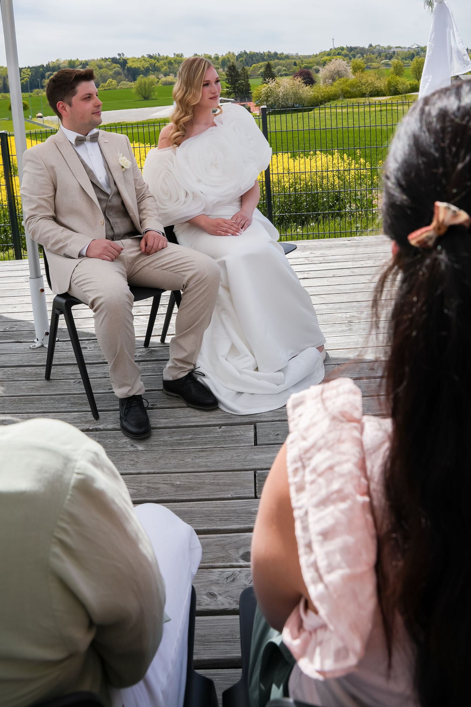 Bride and groom seated at outdoor wedding ceremony on wooden deck with lush green countryside backdrop.