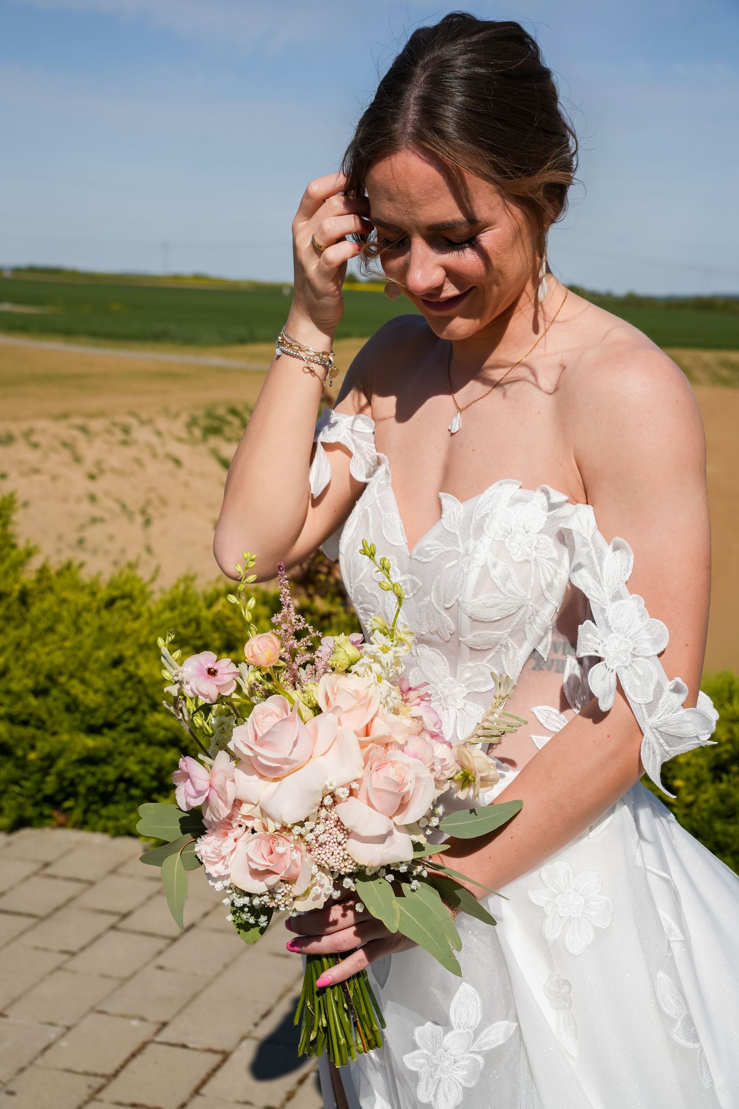 Bride in white lace gown holding a pink rose bouquet, smiling outdoors on a sunny wedding day.