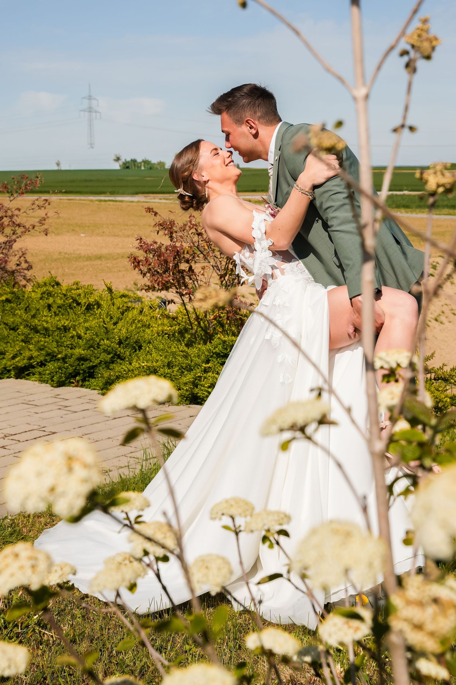 A bride in a white gown and groom in green suit share a romantic kiss outdoors among white flowers and greenery.