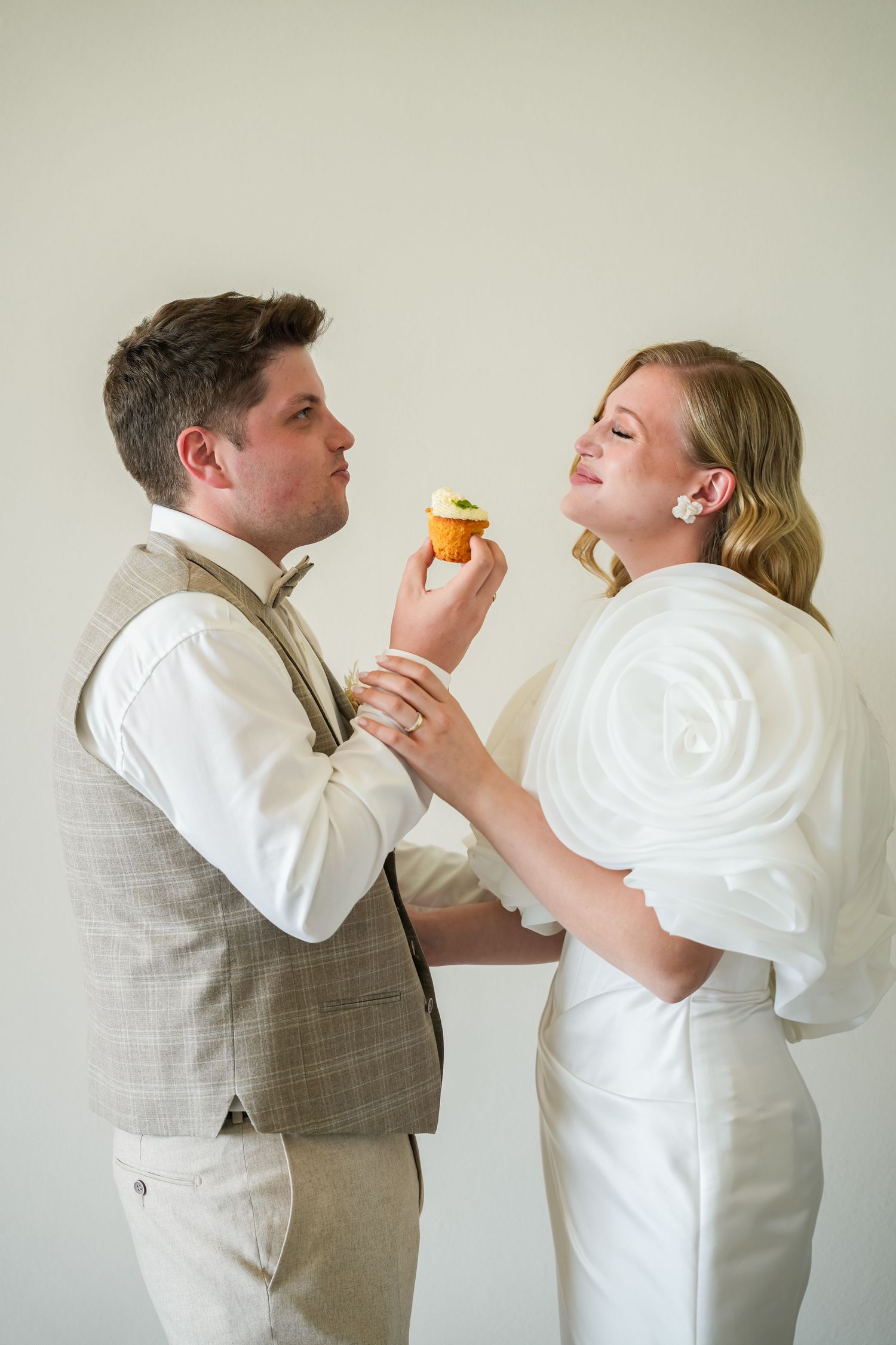 Bride and groom sharing a cupcake, dressed in wedding attire, posing against a light background.