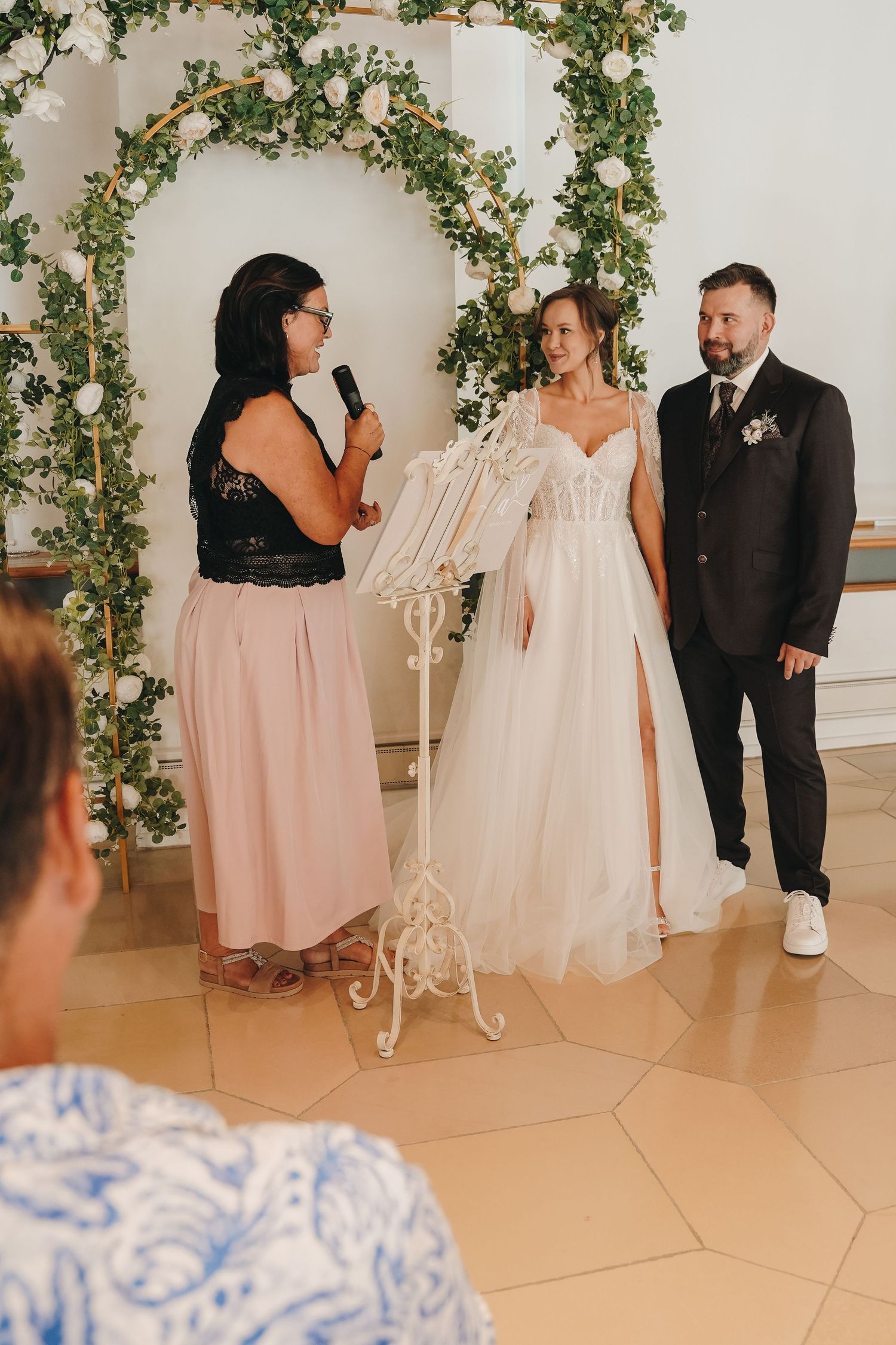 Bride and groom stand at floral arch altar during wedding ceremony while officiant speaks into microphone.