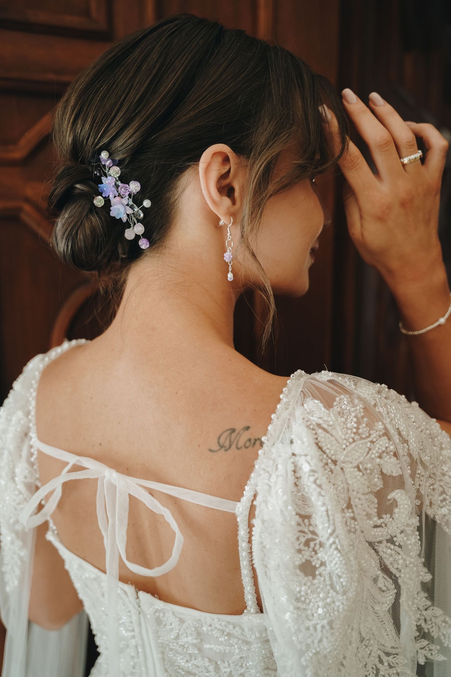 Close-up of bride's back showing lace wedding dress tie, pearl earring, floral hair clip, and neck tattoo.
