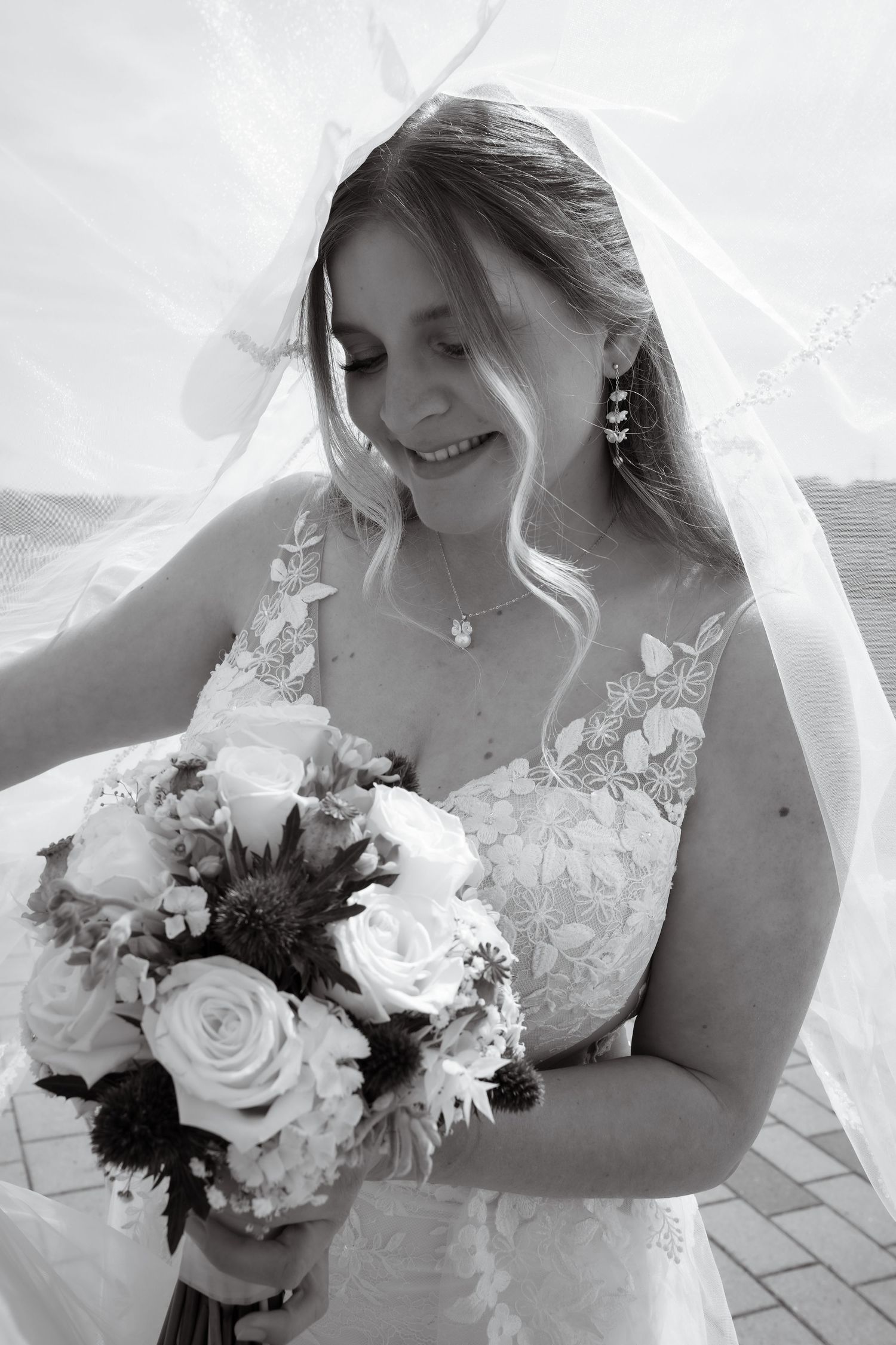 Black and white photo of a smiling bride in lace gown holding a bouquet of roses under her flowing veil.
