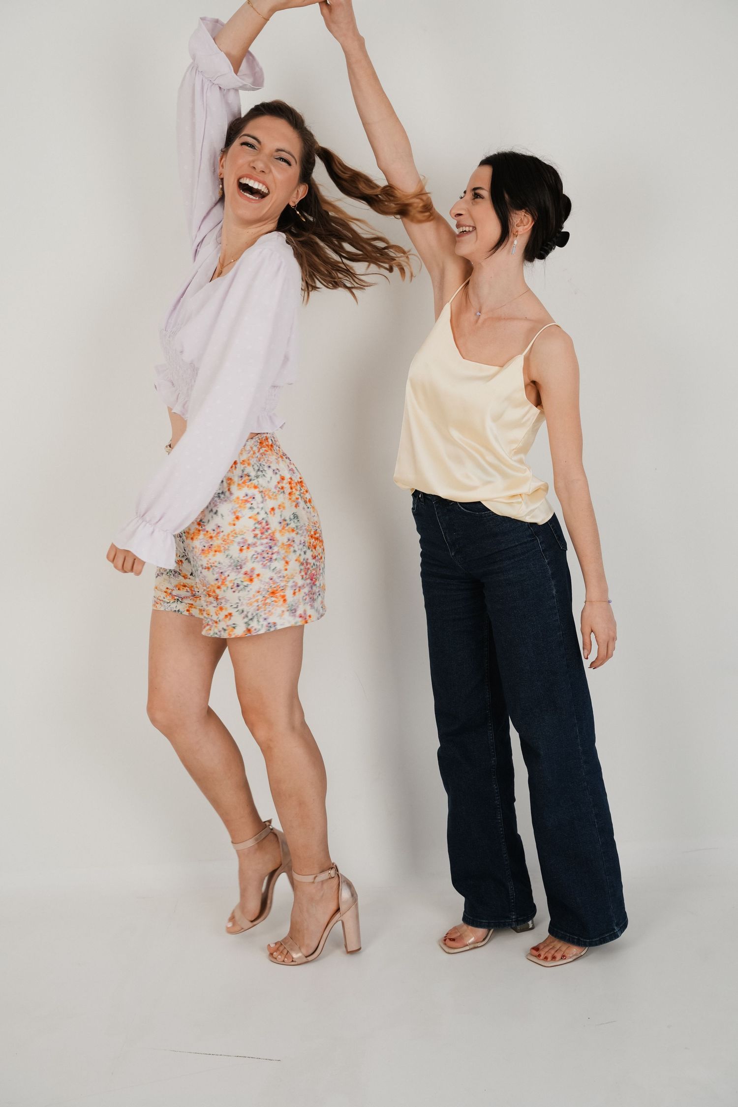 Two women dancing and laughing together, one in floral mini skirt and white top, other in navy wide-leg jeans and yellow cami.