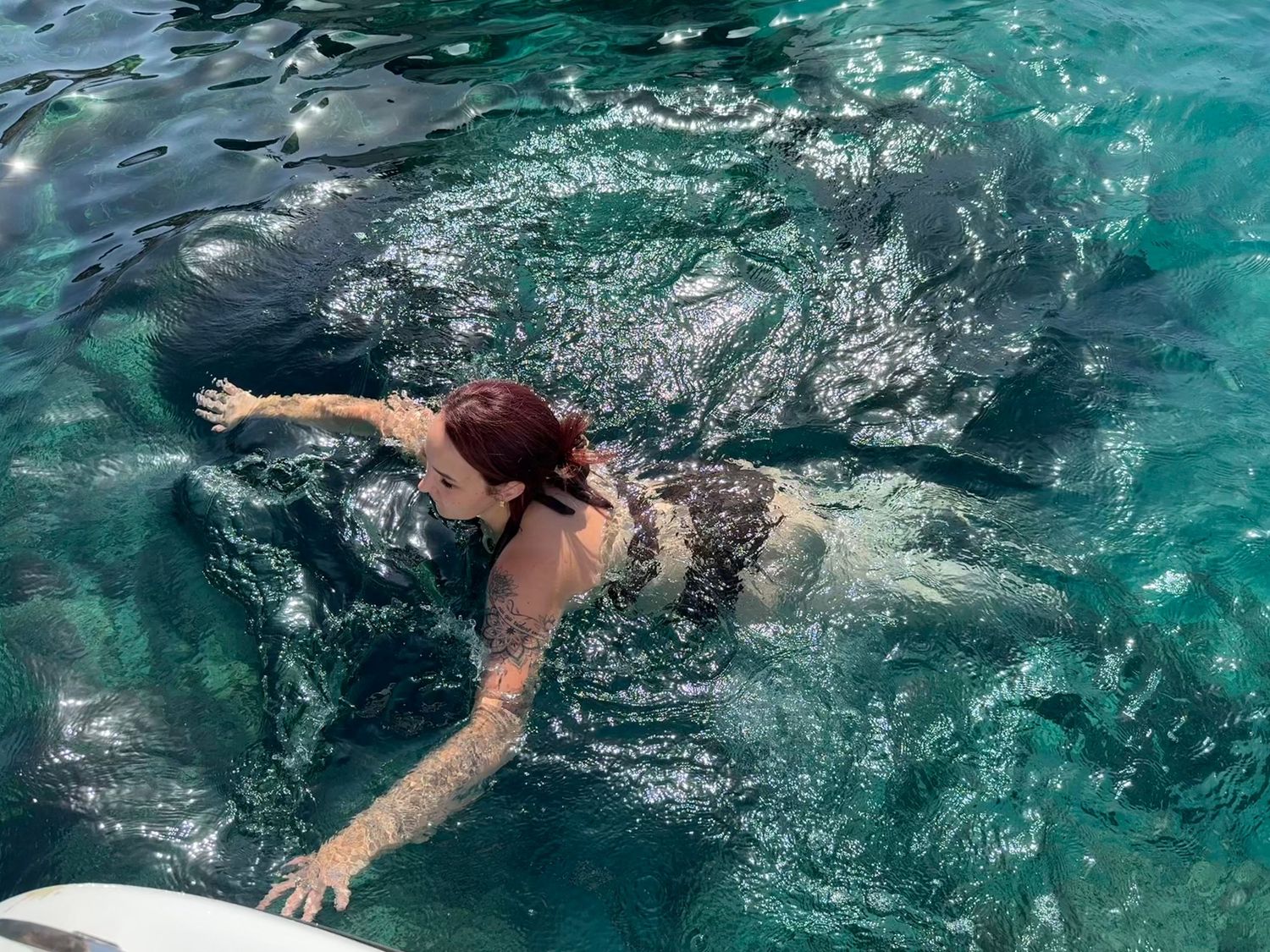 Woman with red hair swimming in clear turquoise ocean water near a white boat on a sunny day.