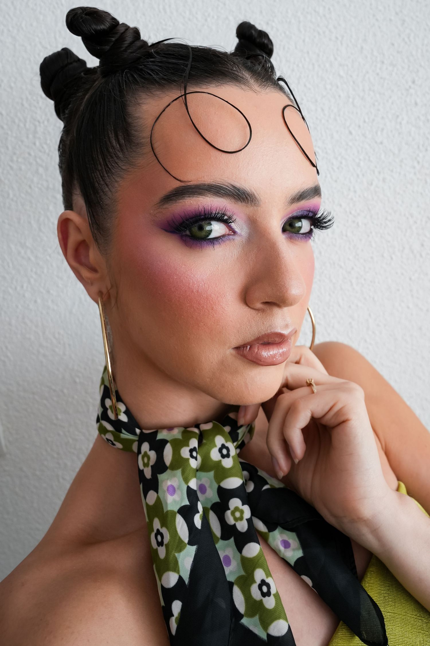 Woman with bold purple eye makeup, space buns, curled face tendrils, floral scarf, and hoop earrings poses against white wall.