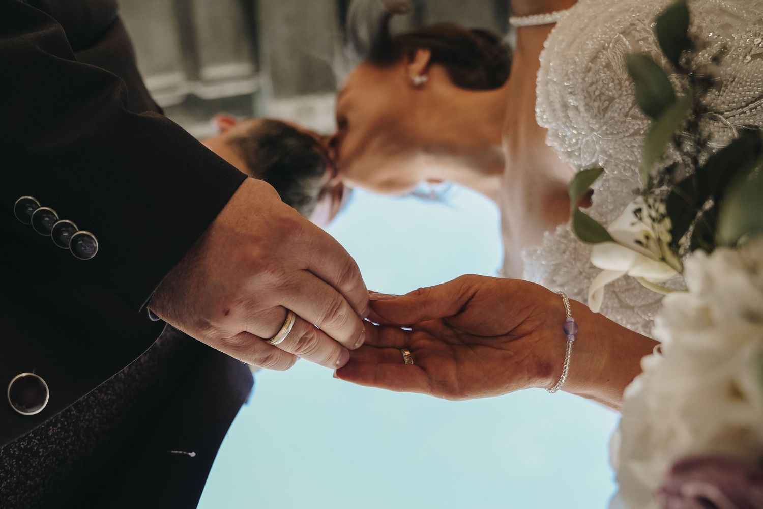 Groom in a dark suit places a wedding ring on the bride's finger during the ceremony, with a white floral bouquet in the foreground.