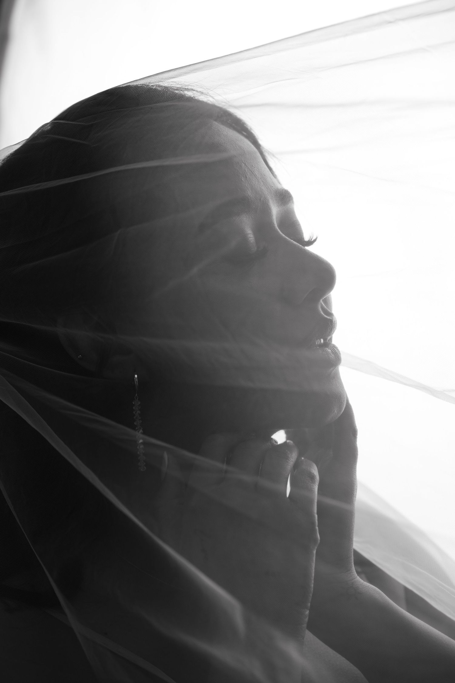 Black and white portrait of a bride with veil draped over her face, looking up in soft backlit light.